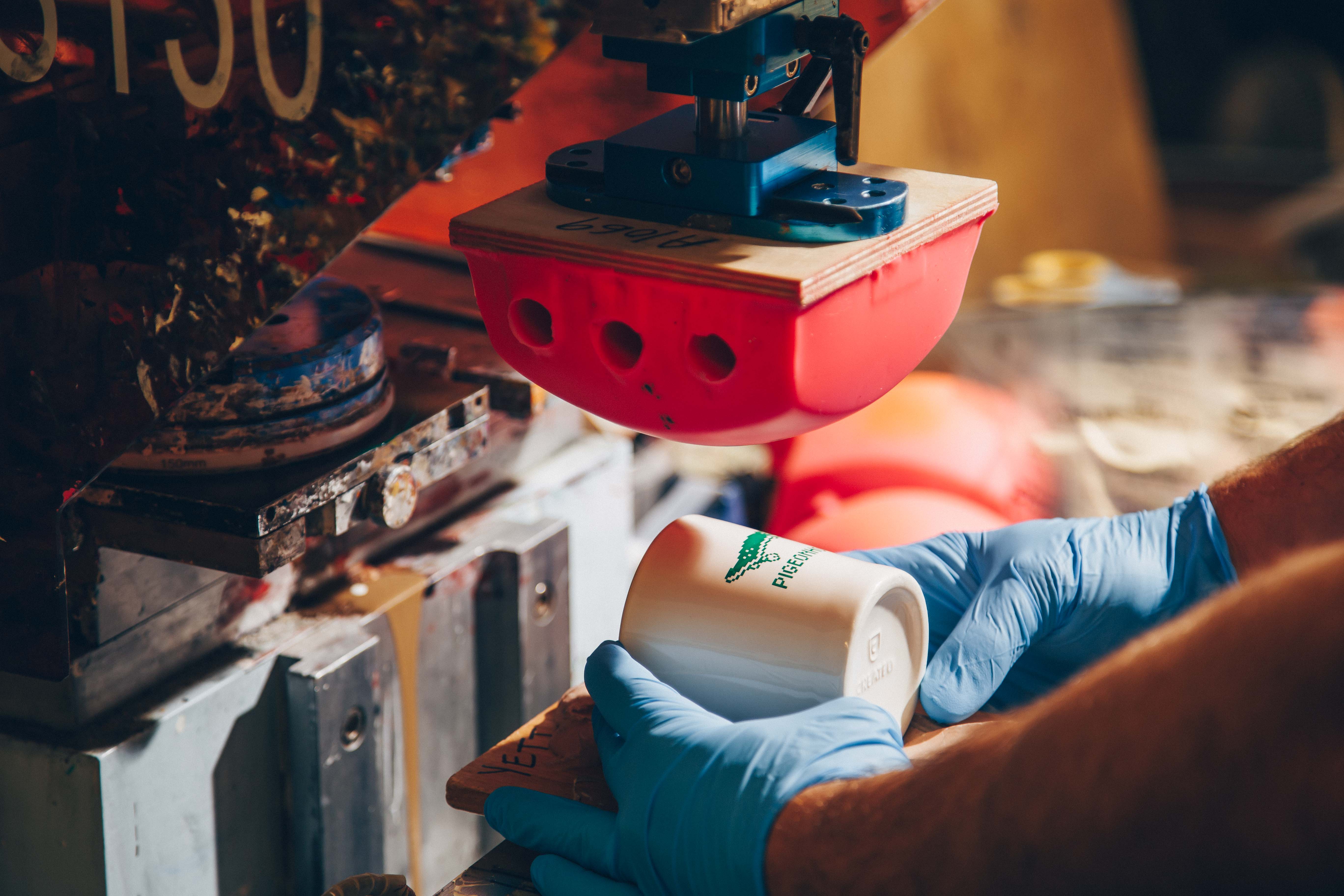 A worker in blue gloves using a pad printing machine to apply a green logo to a white ceramic mug. The machine features a red silicone transfer pad and a well-used print station, showcasing precision in product customization.