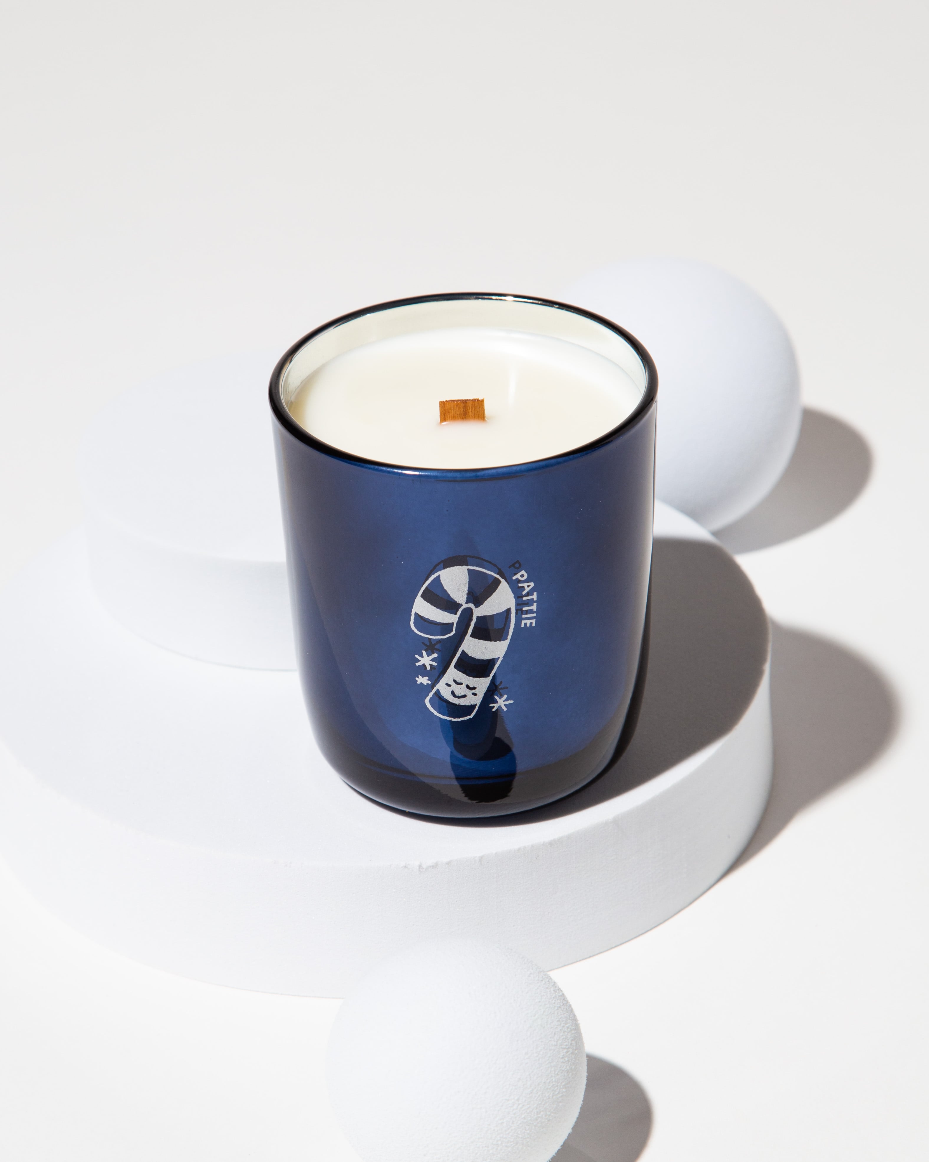 Blue Milk Jar Candle Co branded candle with a wooden wick on a white pedestal, surrounded by white spheres.