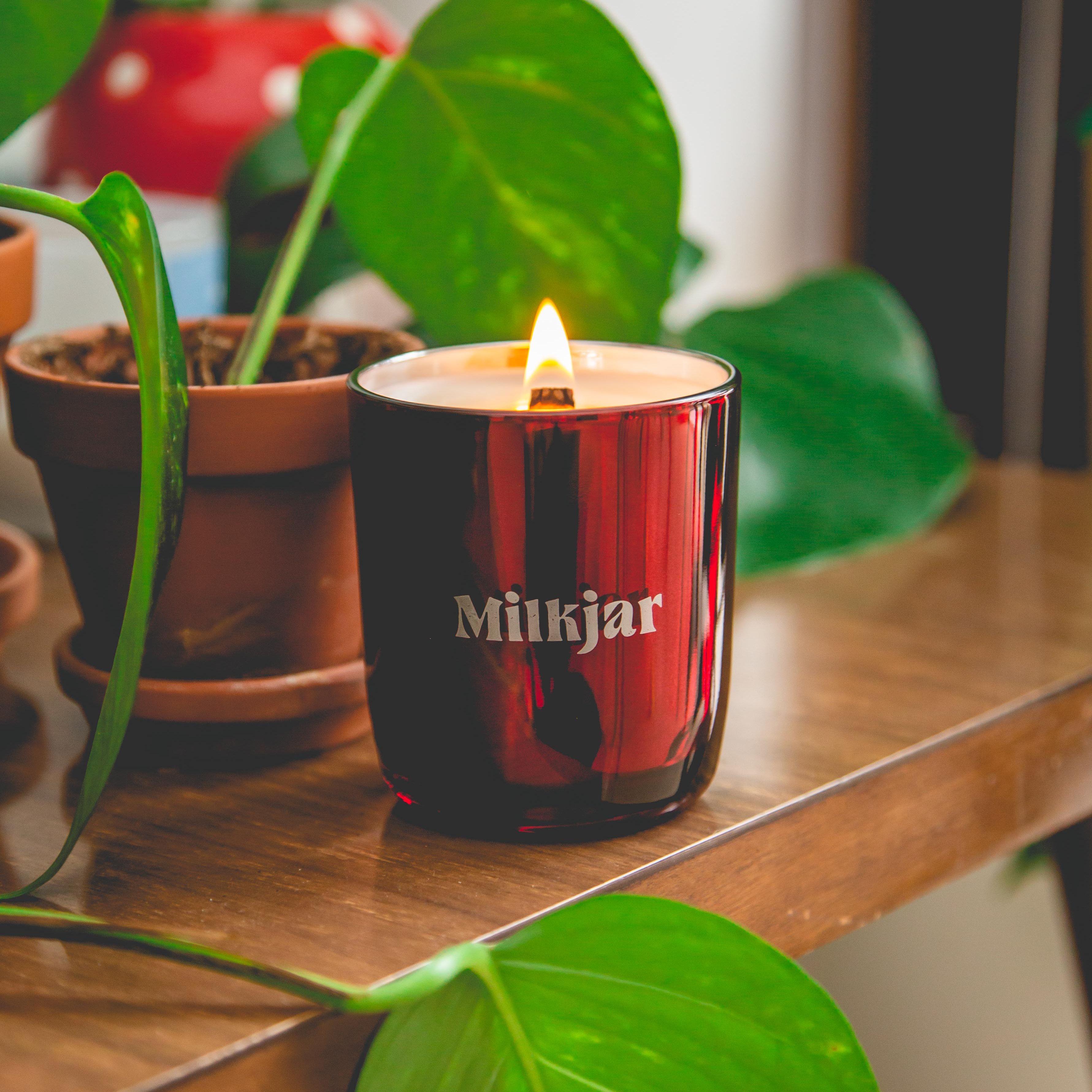 Red candle labeled 'Milkjar' on a wooden table surrounded by green potted plants.