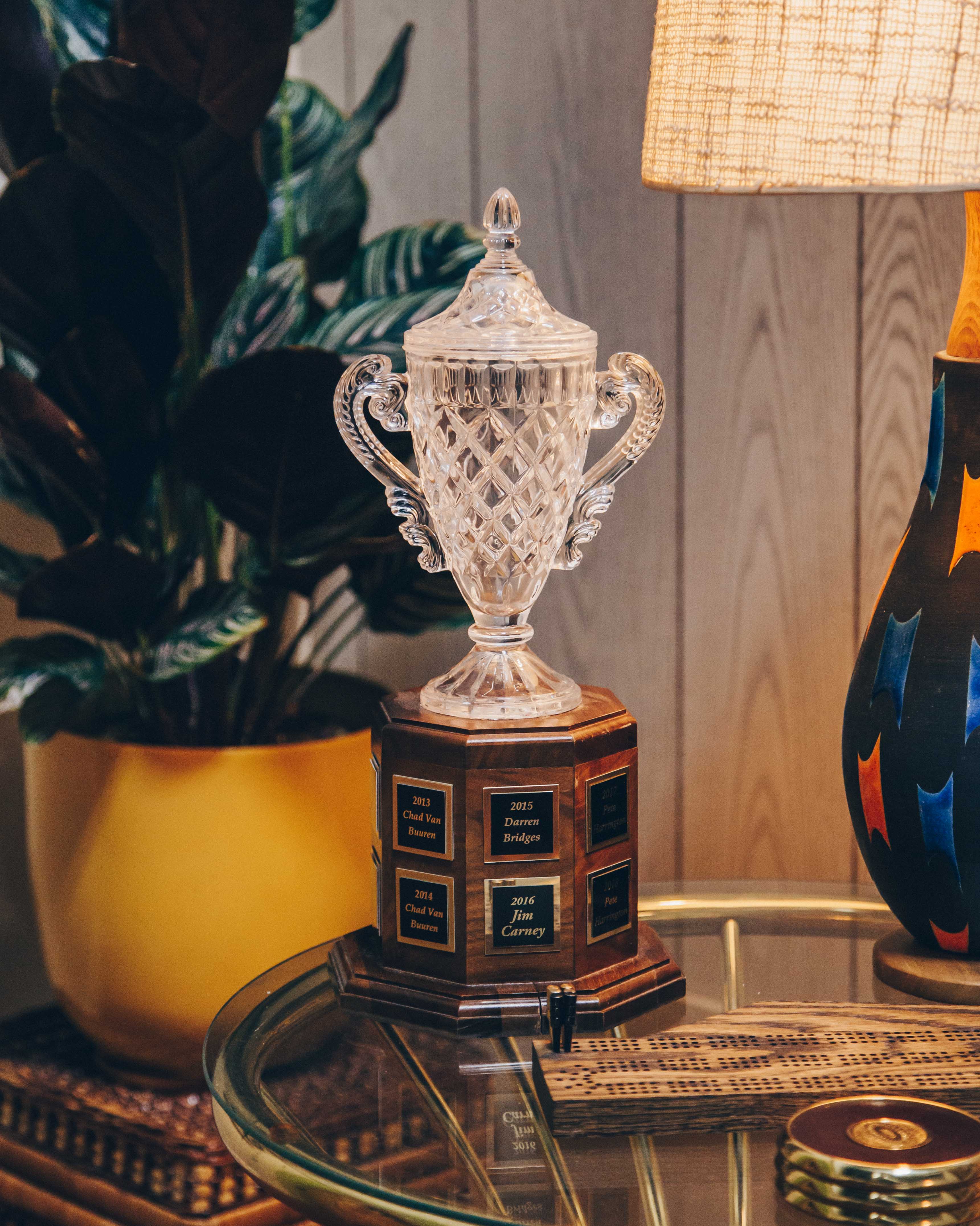 A crystal trophy with ornate handles and a detailed, diamond-cut design. It is placed on a wooden base with engraved plaques listing names and years. The trophy is situated on a glass table, which also holds a wooden cribbage board and a small round object. To the left, there is a large potted plant with dark green leaves in a yellow pot. On the right, a colorful lamp with a textured shade casts a warm glow, creating a cozy and inviting atmosphere. The background consists of wooden paneling, adding to the vintage feel of the setting.