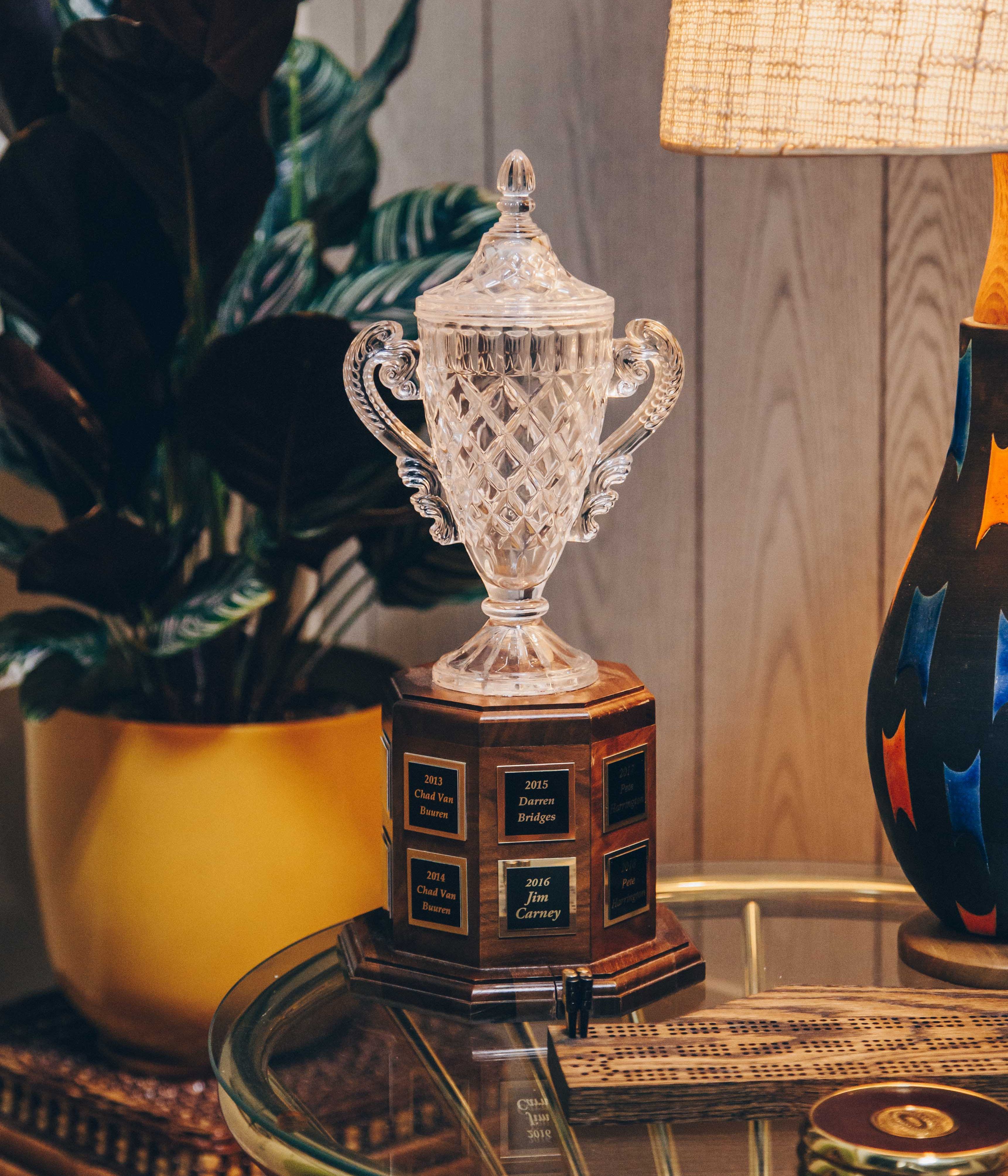 A crystal trophy with ornate handles and a detailed, diamond-cut design. It is placed on a wooden base with engraved plaques listing names and years. The trophy is situated on a glass table, which also holds a wooden cribbage board and a small round object. To the left, there is a large potted plant with dark green leaves in a yellow pot. On the right, a colorful lamp with a textured shade casts a warm glow, creating a cozy and inviting atmosphere. The background consists of wooden paneling, adding to the vintage feel of the setting.