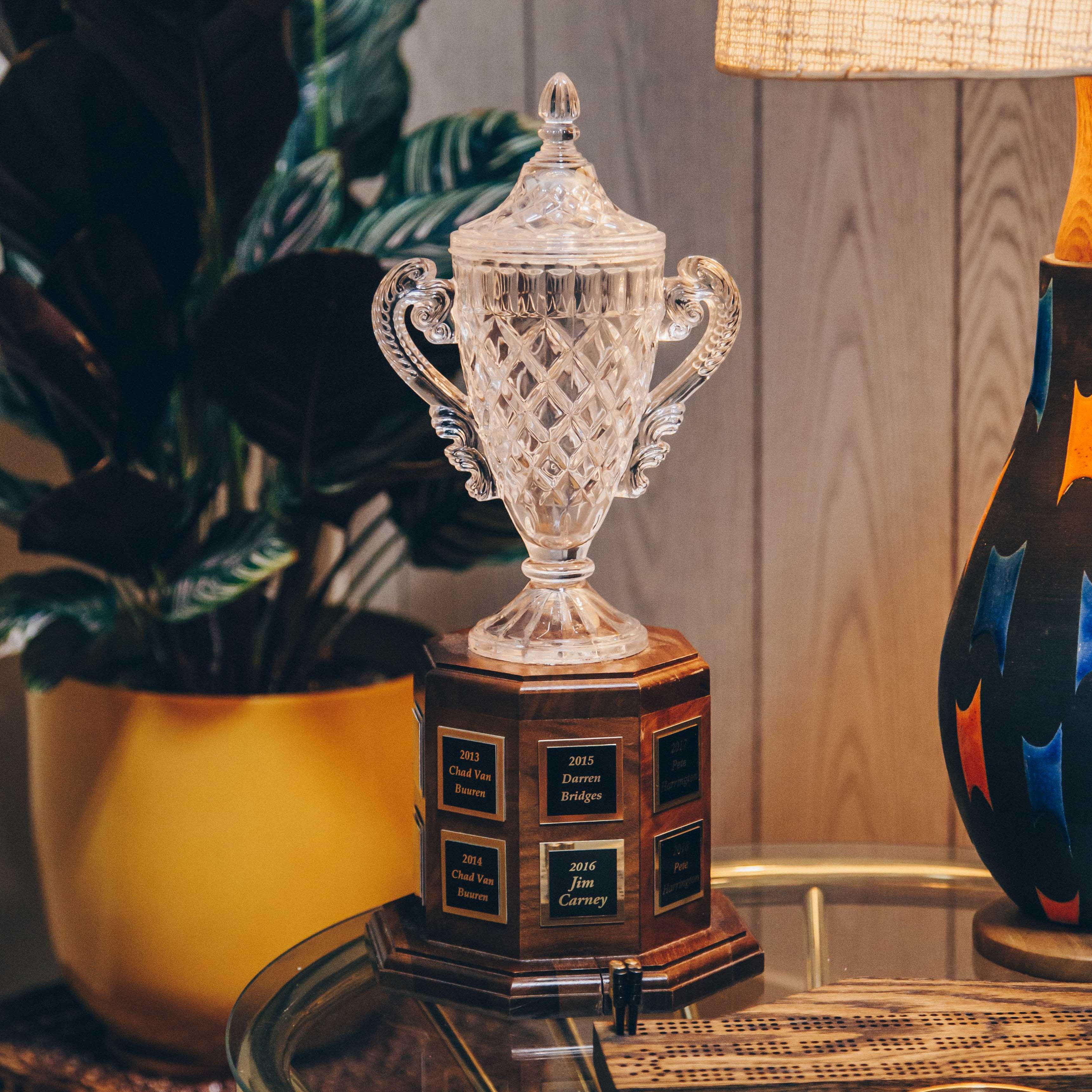 A crystal trophy with ornate handles and a detailed, diamond-cut design. It is placed on a wooden base with engraved plaques listing names and years. The trophy is situated on a glass table, which also holds a wooden cribbage board and a small round object. To the left, there is a large potted plant with dark green leaves in a yellow pot. On the right, a colorful lamp with a textured shade casts a warm glow, creating a cozy and inviting atmosphere. The background consists of wooden paneling, adding to the vintage feel of the setting.