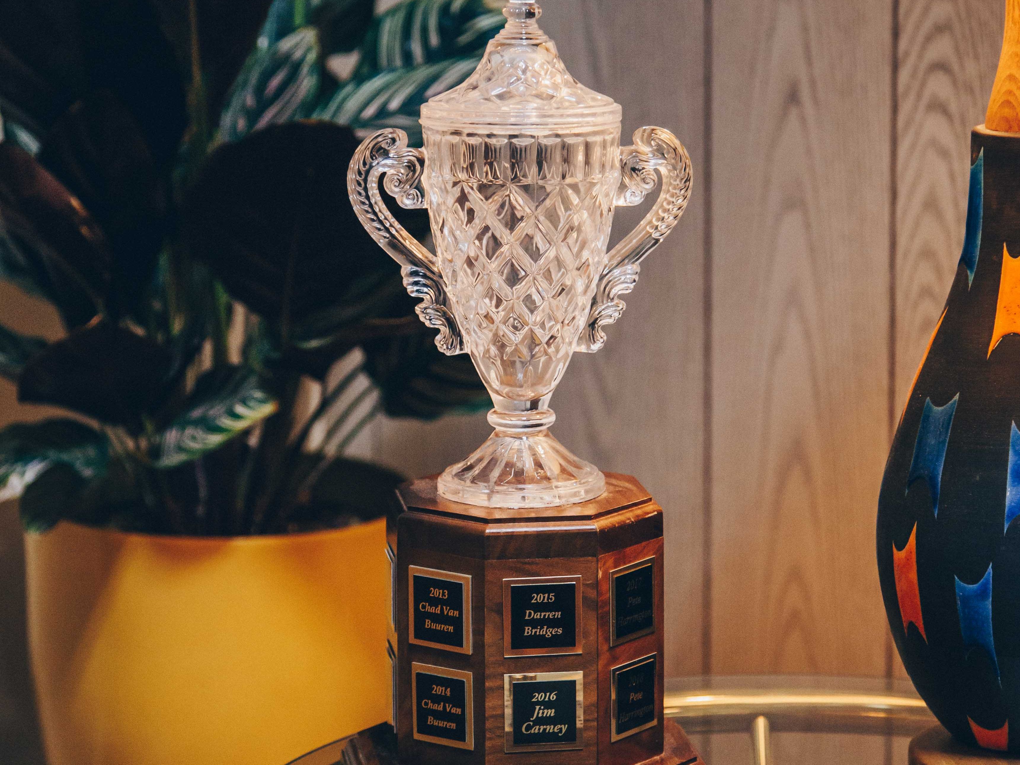 A crystal trophy with ornate handles and a detailed, diamond-cut design. It is placed on a wooden base with engraved plaques listing names and years. The trophy is situated on a glass table, which also holds a wooden cribbage board and a small round object. To the left, there is a large potted plant with dark green leaves in a yellow pot. On the right, a colorful lamp with a textured shade casts a warm glow, creating a cozy and inviting atmosphere. The background consists of wooden paneling, adding to the vintage feel of the setting.