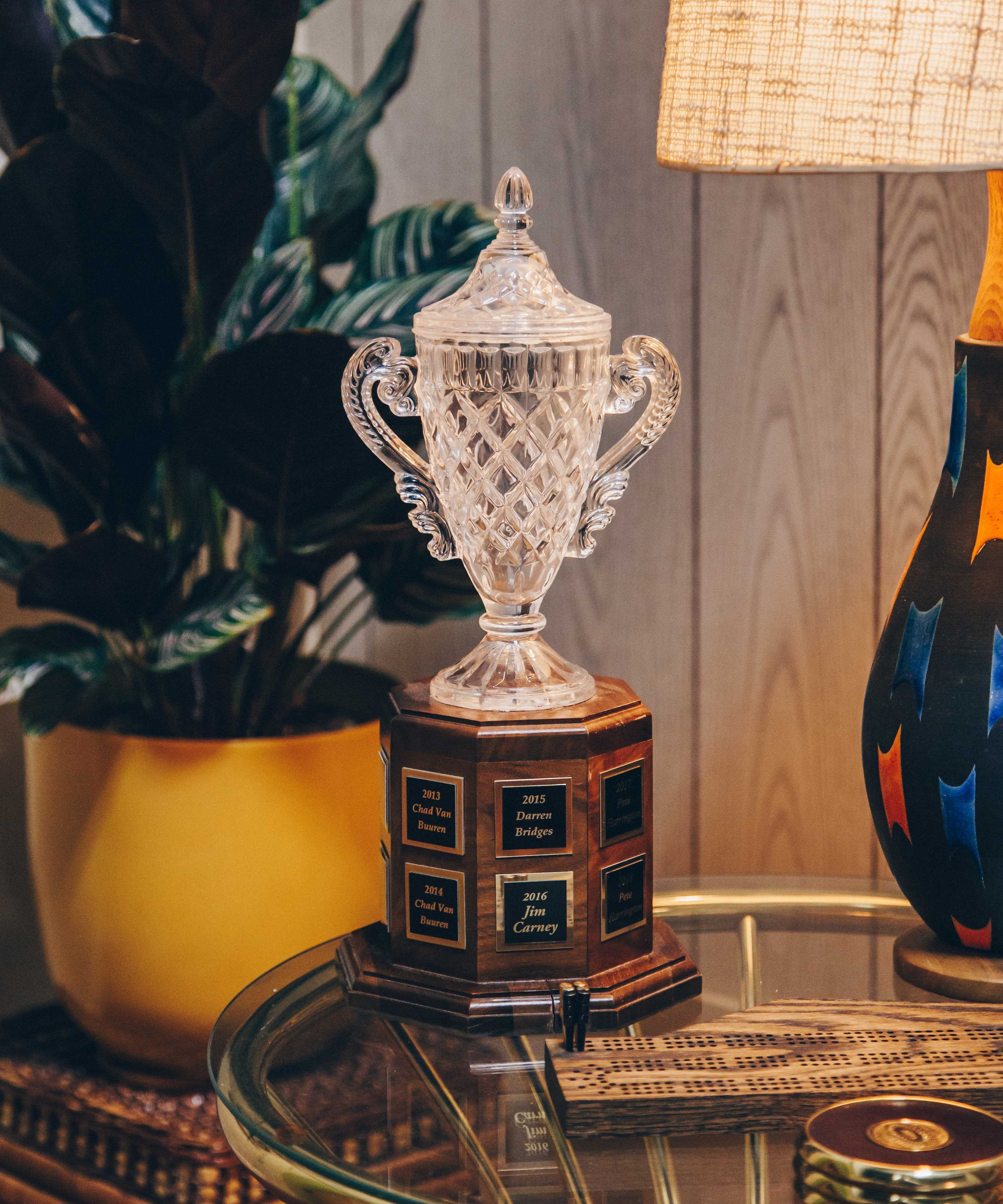 A crystal trophy with ornate handles and a detailed, diamond-cut design. It is placed on a wooden base with engraved plaques listing names and years. The trophy is situated on a glass table, which also holds a wooden cribbage board and a small round object. To the left, there is a large potted plant with dark green leaves in a yellow pot. On the right, a colorful lamp with a textured shade casts a warm glow, creating a cozy and inviting atmosphere. The background consists of wooden paneling, adding to the vintage feel of the setting.