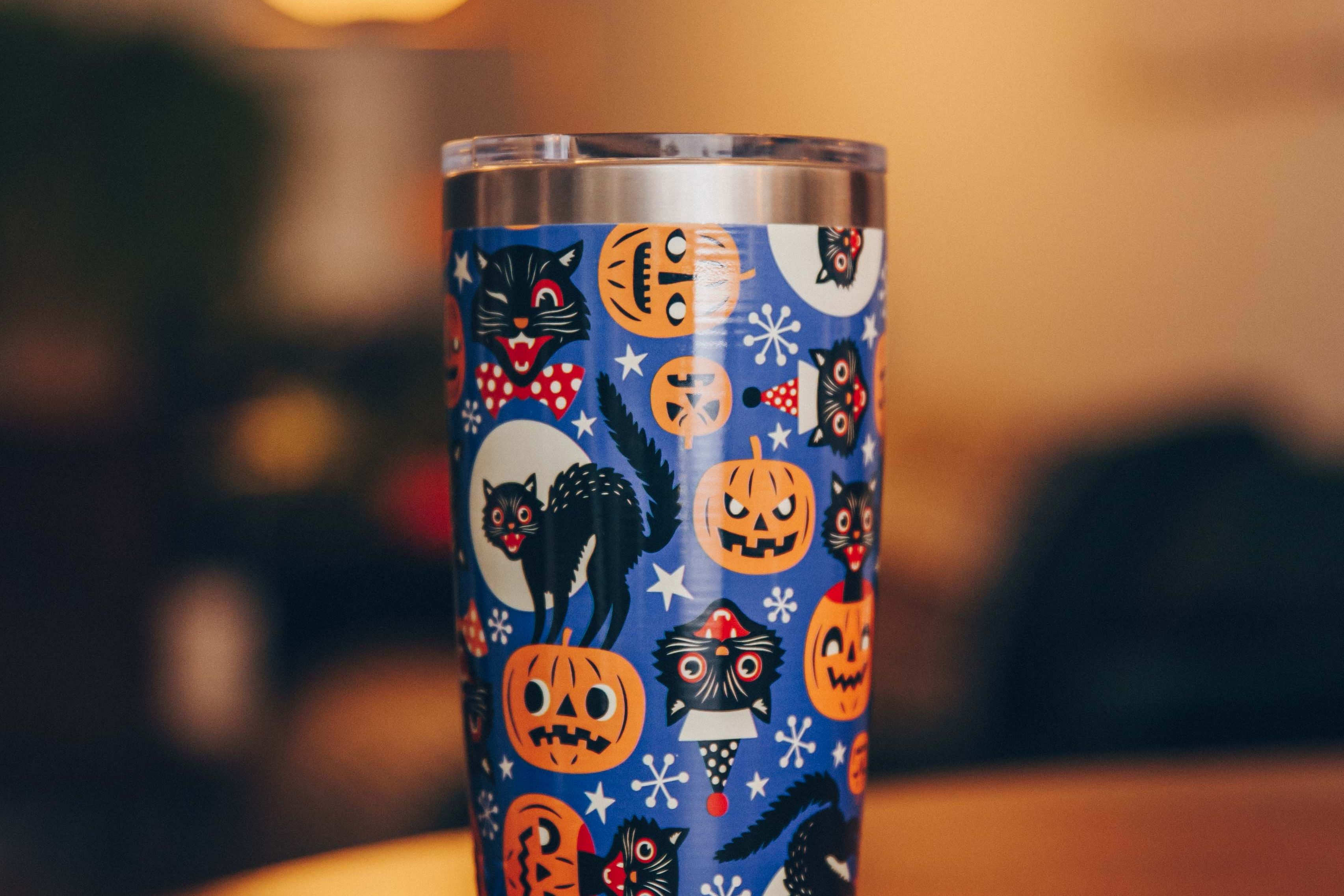 Stainless steel tumbler with Halloween design featuring black cats and pumpkins on a table.