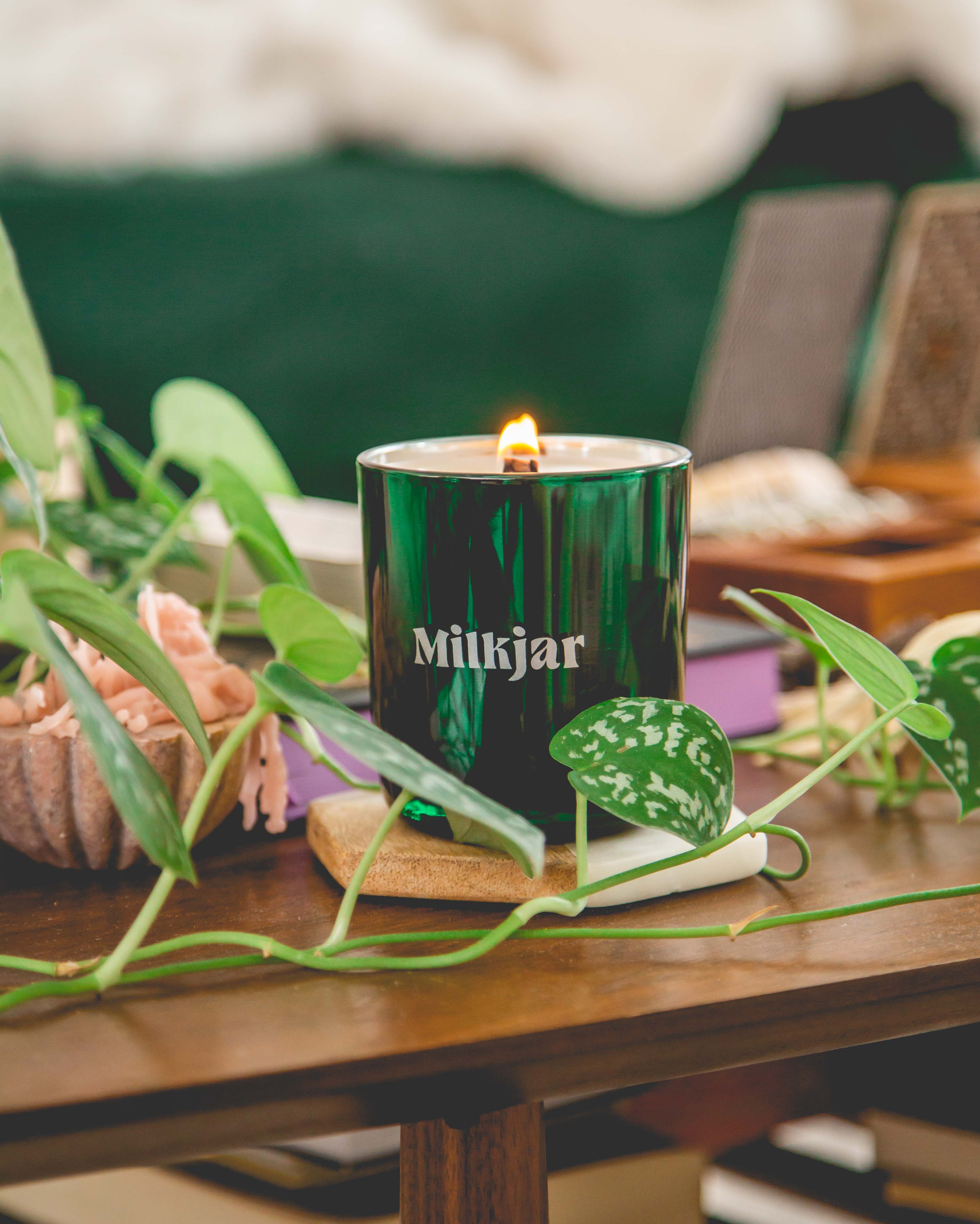 Green candle labeled 'Milkjar' on a wooden table surrounded by green leaves and decor.