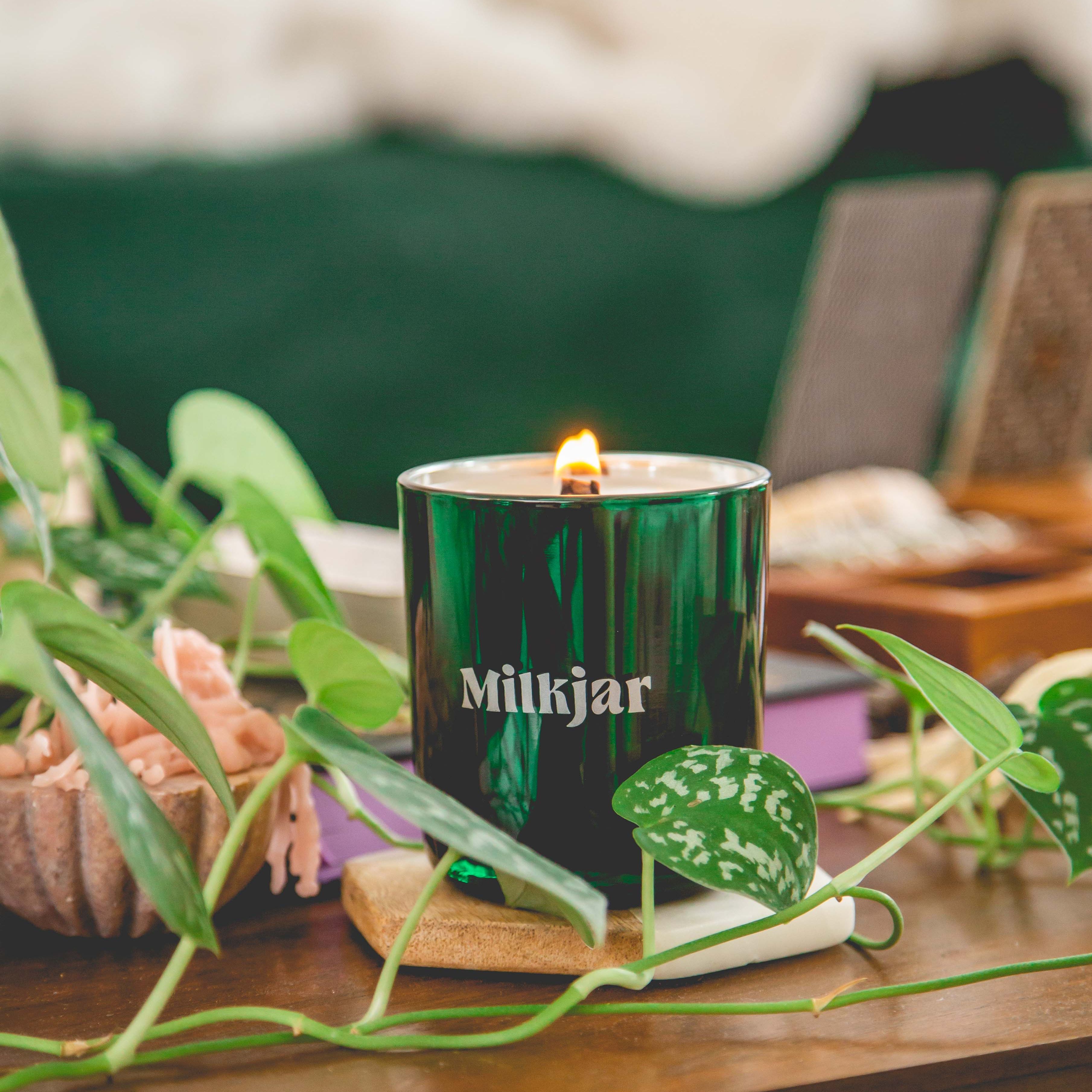 Green candle labeled 'Milkjar' on a wooden table surrounded by green leaves and decor.