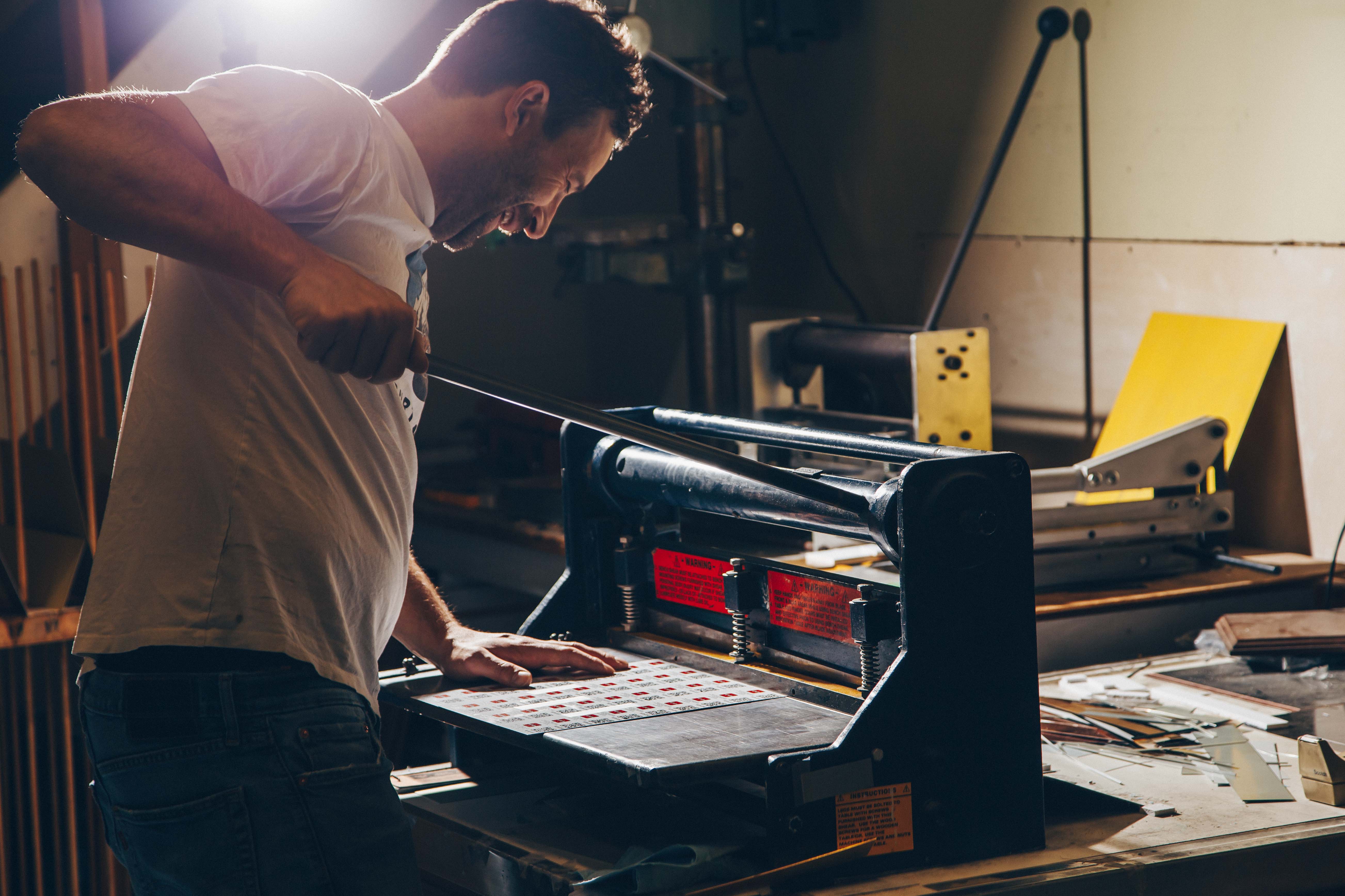 A man operating a manual cutting press in a workshop. He is wearing a white t-shirt and jeans, and is focused on the task at hand, using both hands to manipulate the press. The workspace is cluttered with various tools and materials, suggesting an environment of creativity and craftsmanship.