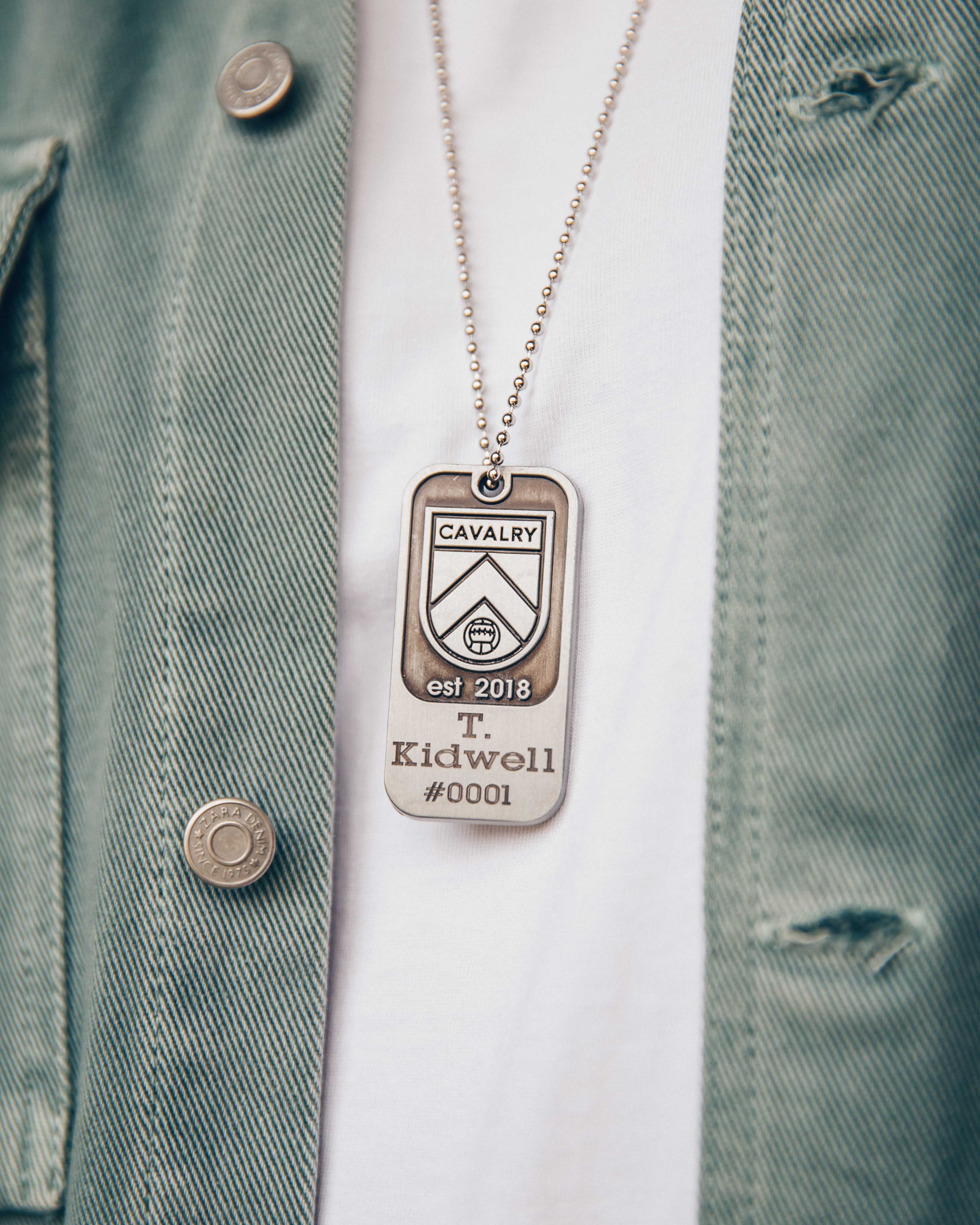 Close-up of a Cavalry dog tag necklace on a white shirt and green jacket.