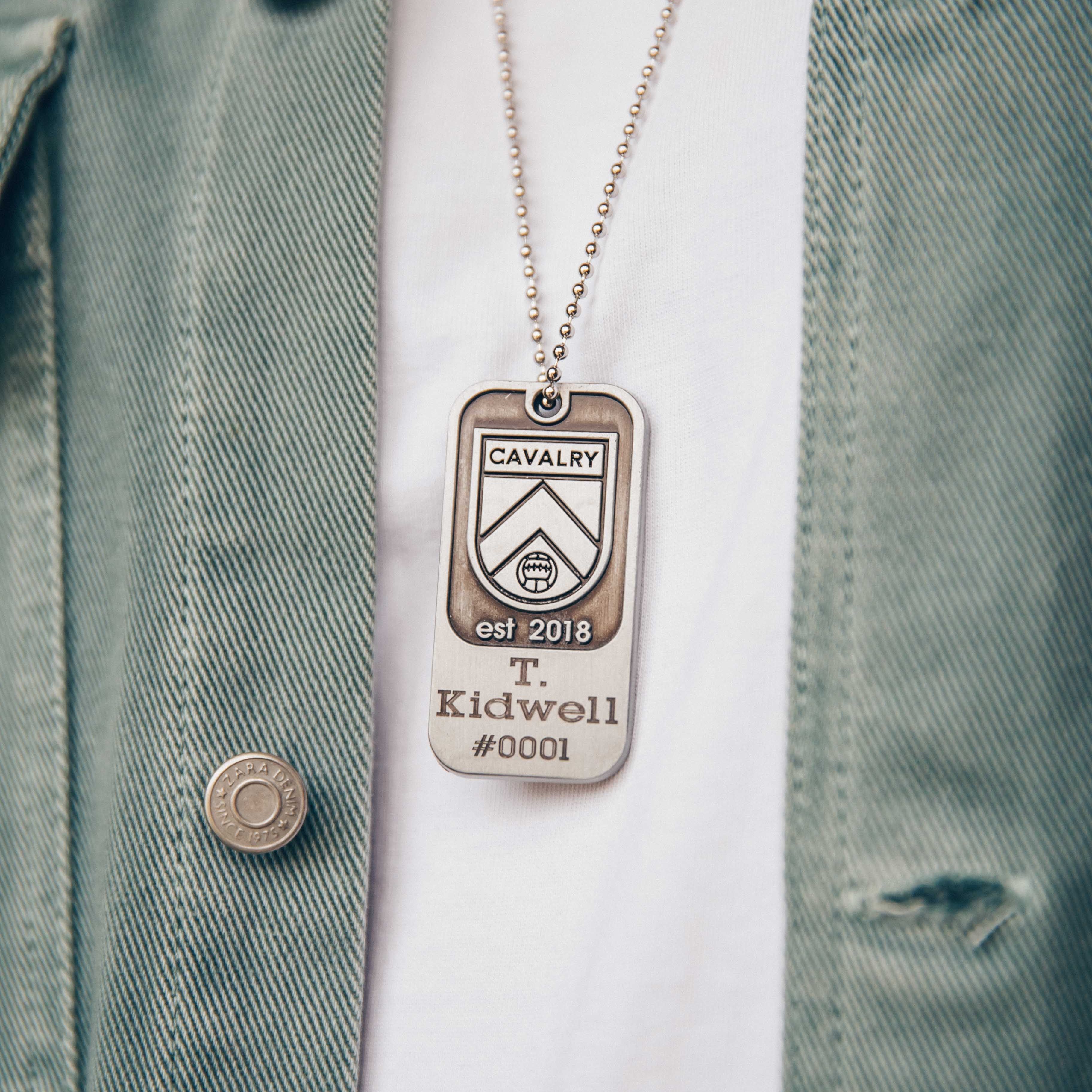 Close-up of a Cavalry dog tag necklace on a white shirt and green jacket.