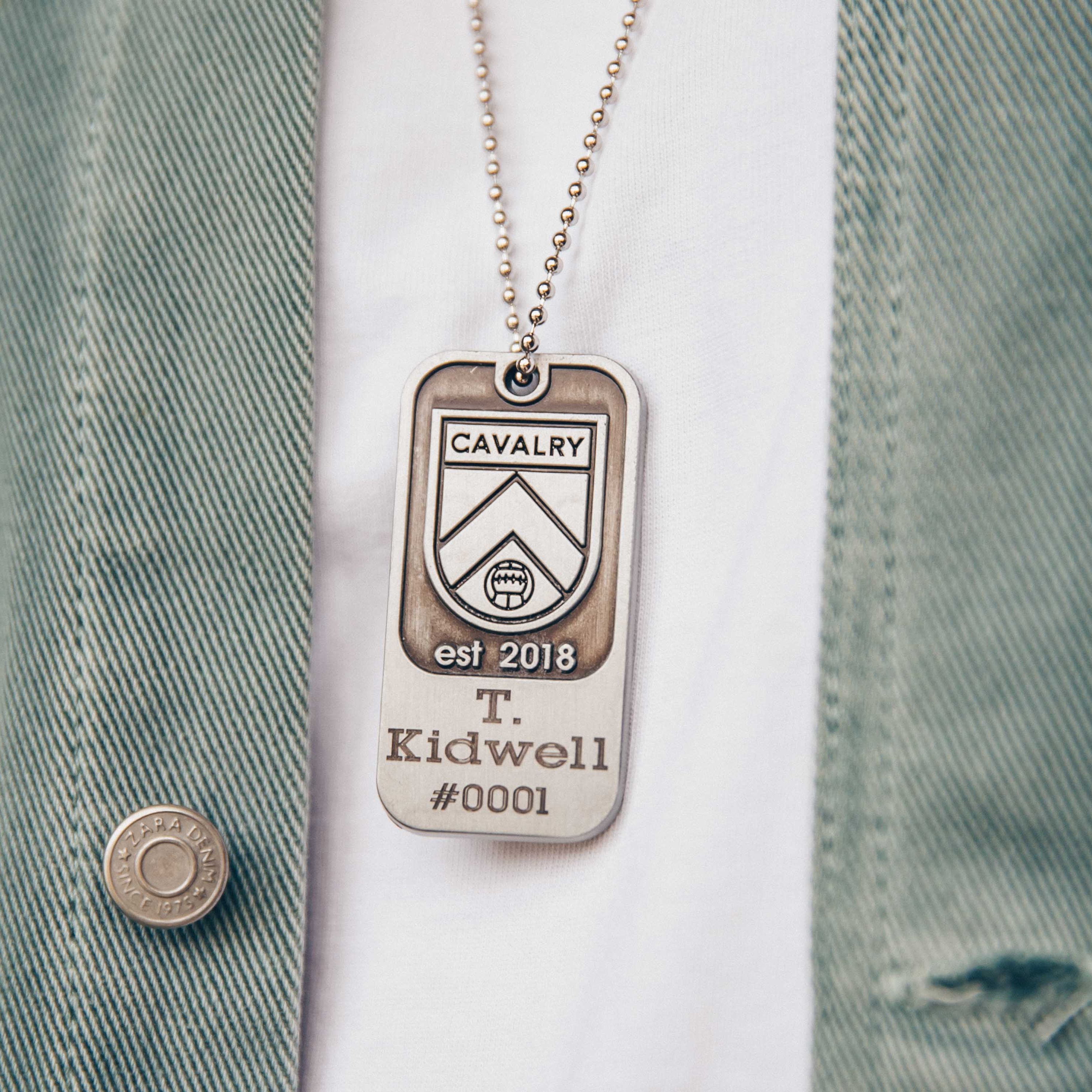 Close-up of a Cavalry dog tag necklace on a white shirt and green jacket.