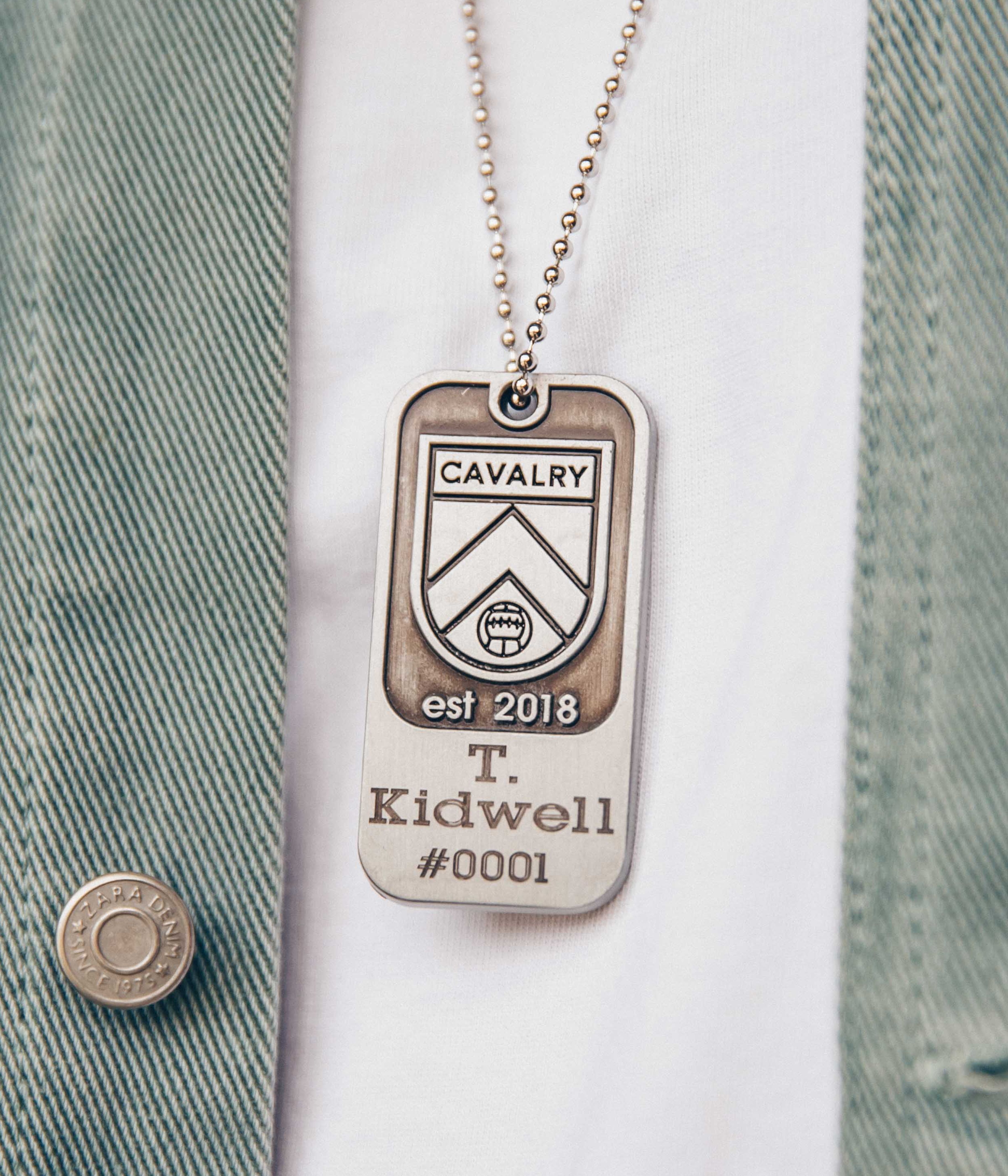 Close-up of a Cavalry dog tag necklace on a white shirt and green jacket.