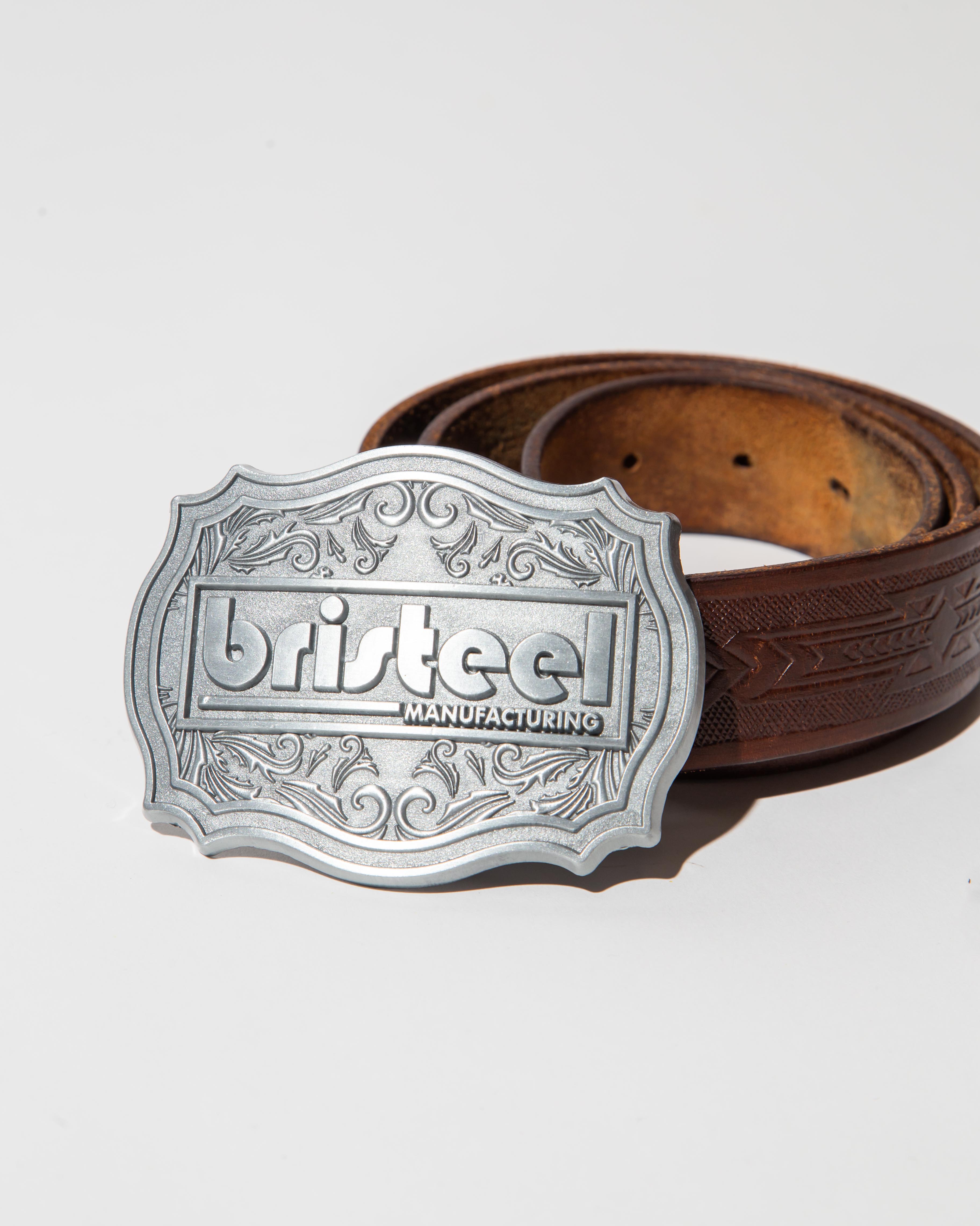 A belt with a prominent metallic buckle. The buckle is intricately designed with ornate patterns and the word 'bristeel' embossed in the center, along with the word 'MANUFACTURING' below it. The buckle has a silver finish with a decorative border. The belt itself is made of brown leather, featuring embossed patterns that add texture and detail. The lighting is bright and direct, casting a shadow of the buckle onto the surface, which is a plain, light-colored background.