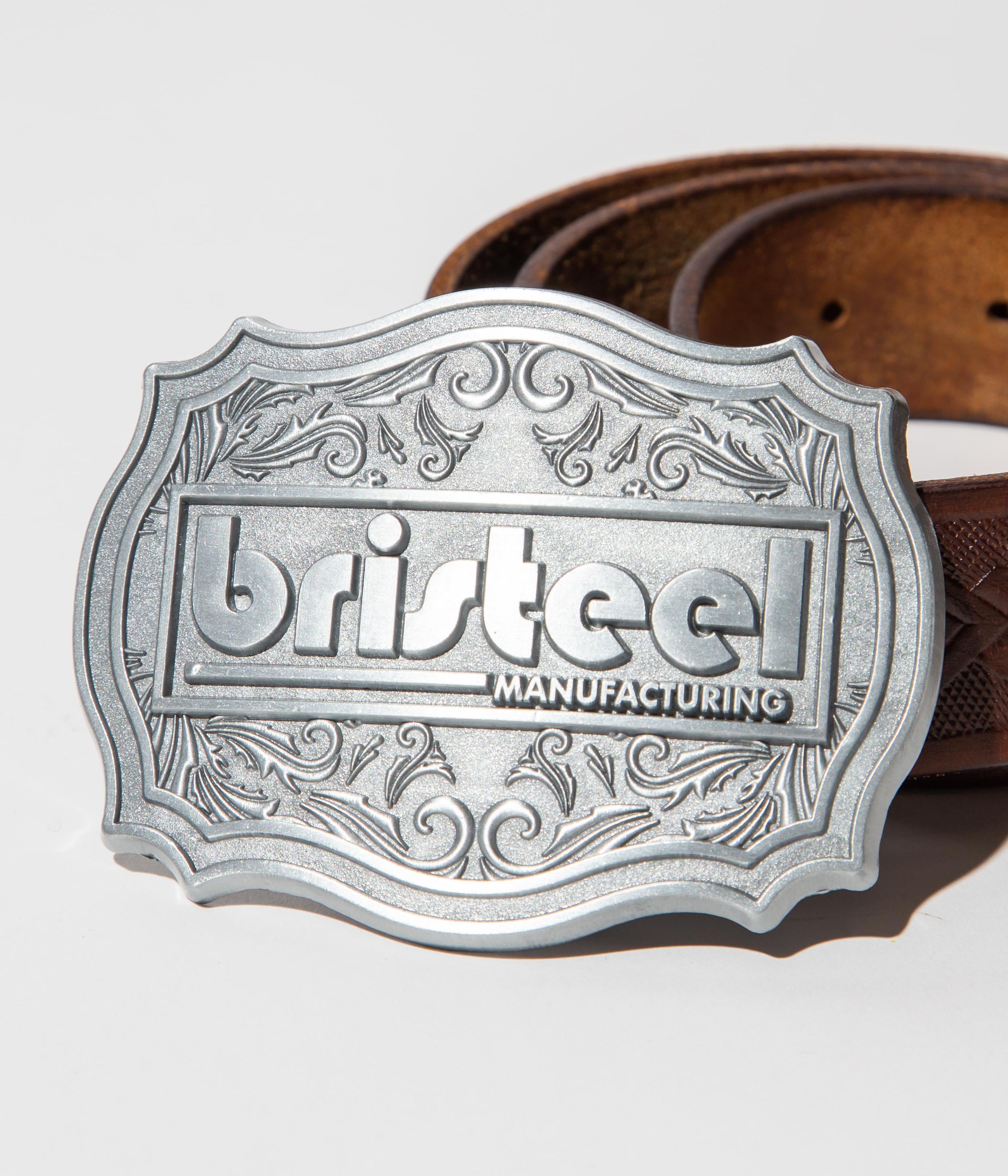 A belt with a prominent metallic buckle. The buckle is intricately designed with ornate patterns and the word 'bristeel' embossed in the center, along with the word 'MANUFACTURING' below it. The buckle has a silver finish with a decorative border. The belt itself is made of brown leather, featuring embossed patterns that add texture and detail. The lighting is bright and direct, casting a shadow of the buckle onto the surface, which is a plain, light-colored background.