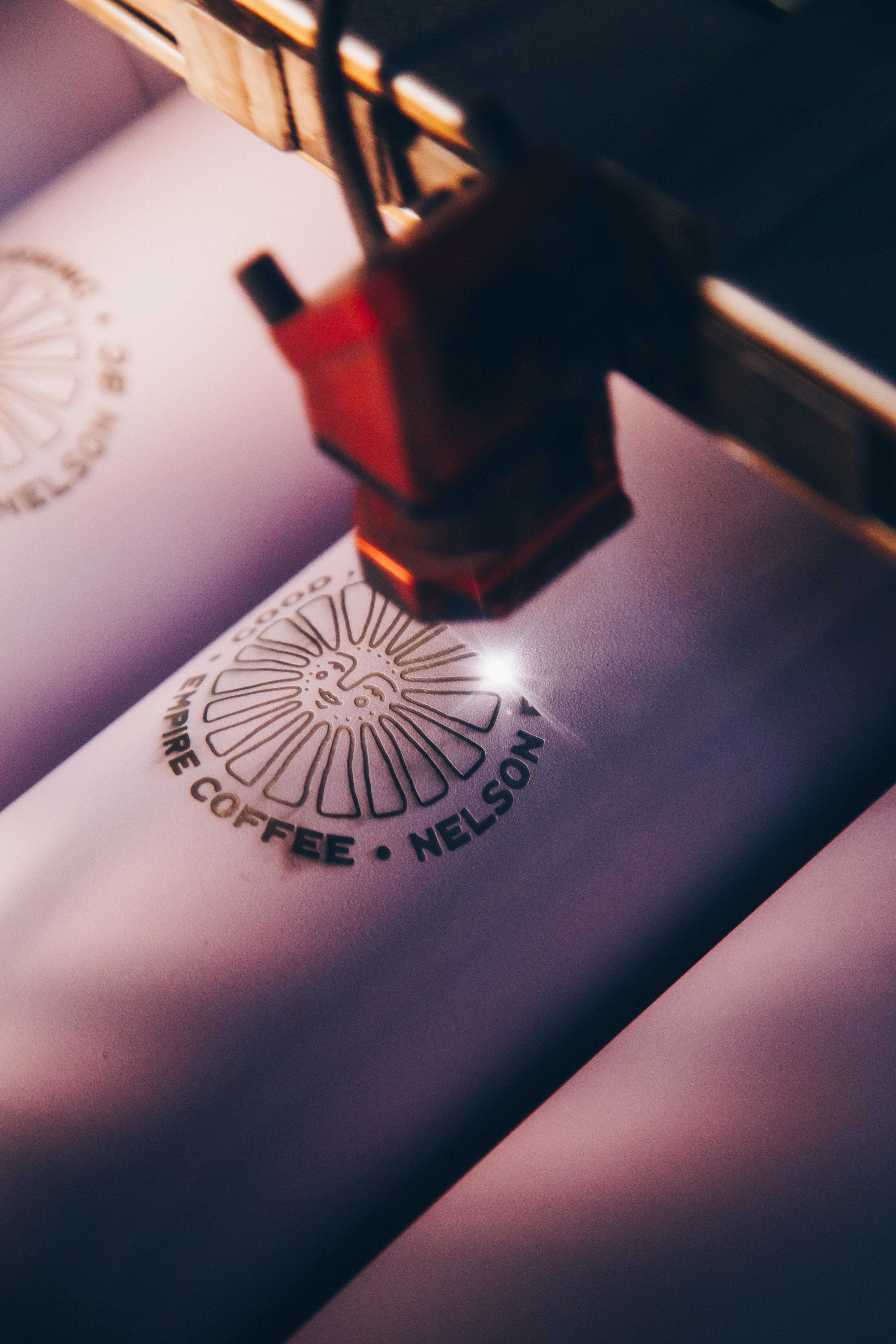 A close-up of a laser engraving machine etching the "Good Morning Coffee, Nelson BC" logo onto white drinkware. The precise beam highlights the customization process for high-quality branded tumblers.