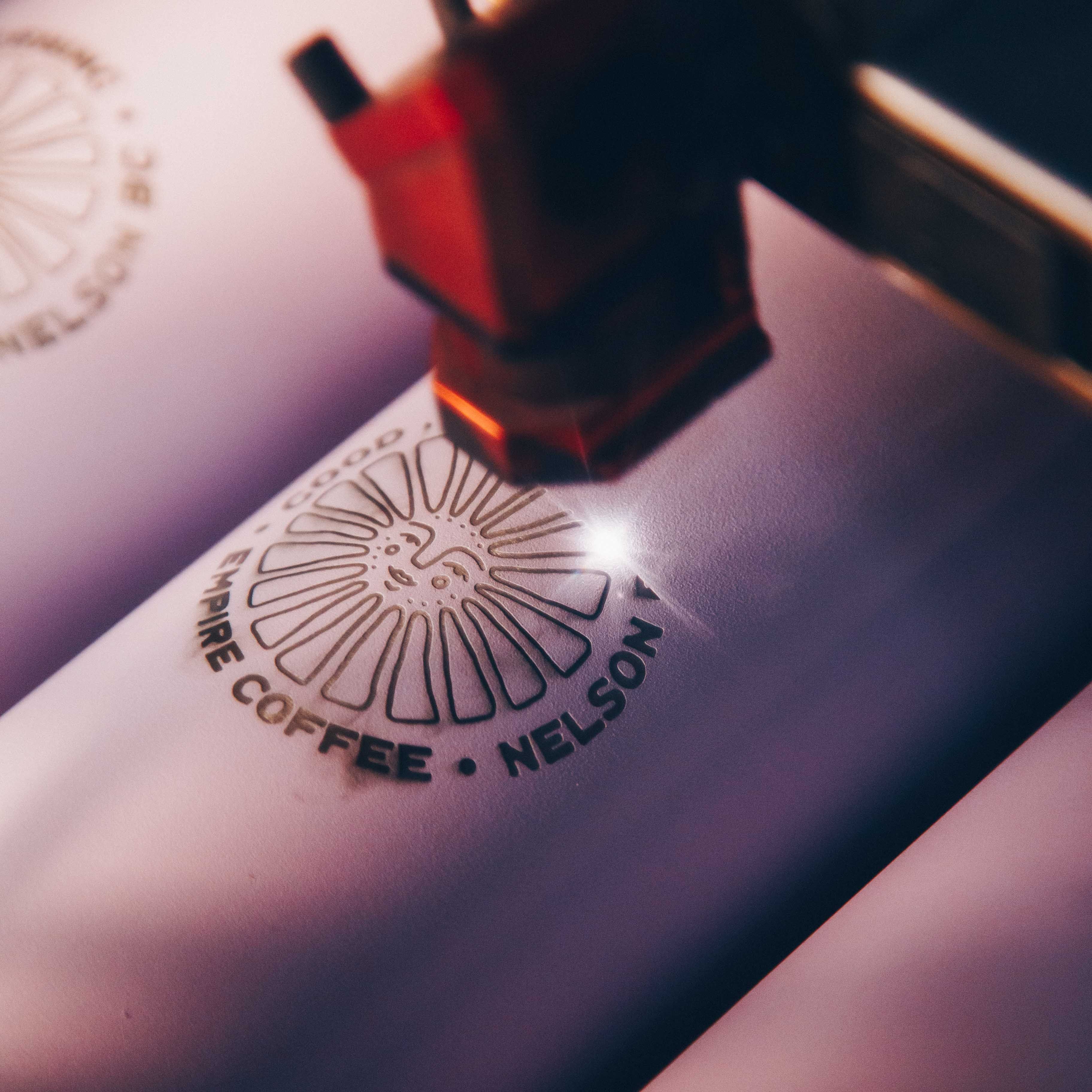 A close-up of a laser engraving machine etching the "Good Morning Coffee, Nelson BC" logo onto white drinkware. The precise beam highlights the customization process for high-quality branded tumblers.