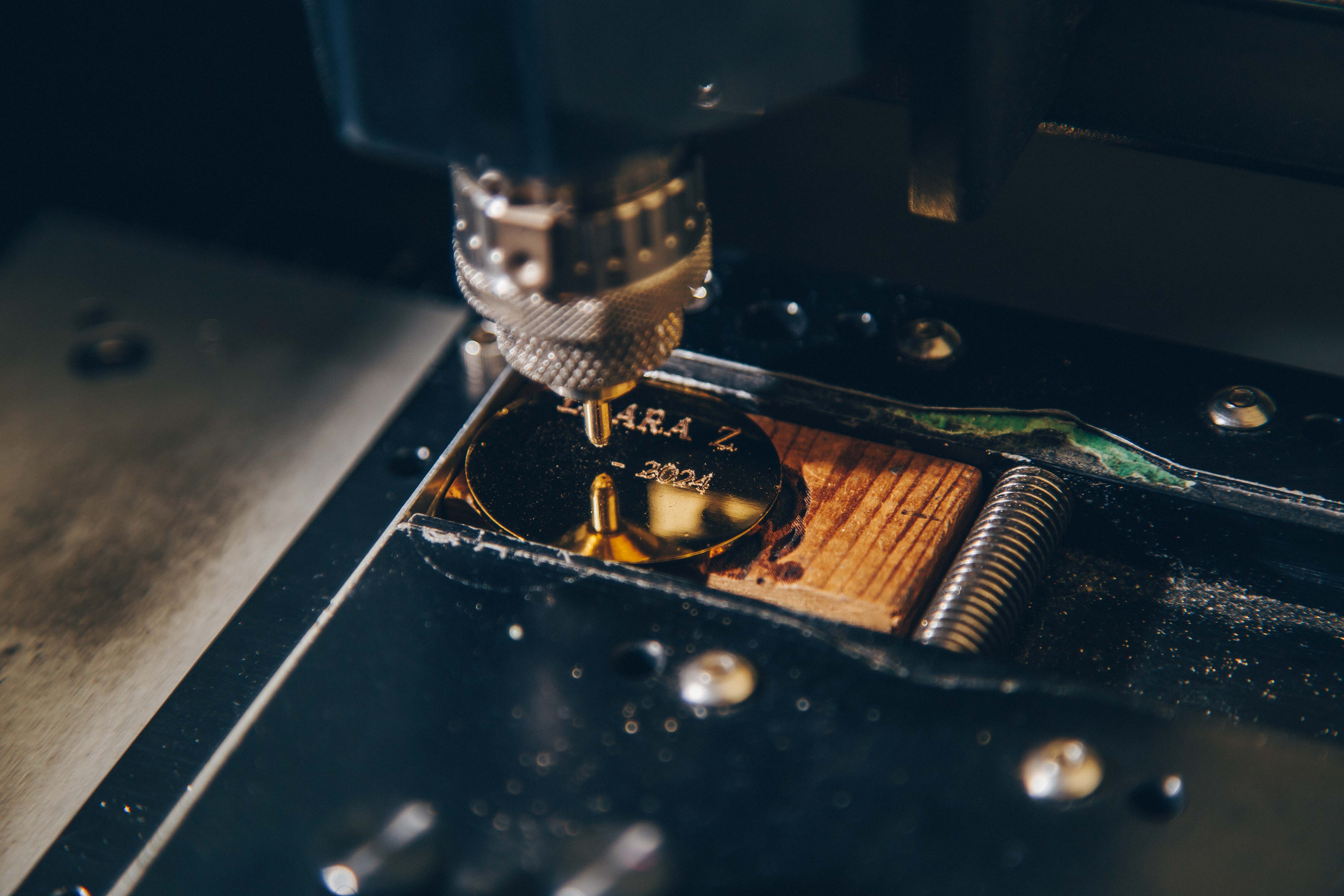 A close-up view of a metal engraving process. A precision engraving machine is actively etching text onto a shiny, circular metal plate. The text 'ARA Z' and '2024' is visible on the surface of the plate. The machine's engraving tool is positioned directly above the plate, with its metallic, textured surface in focus.