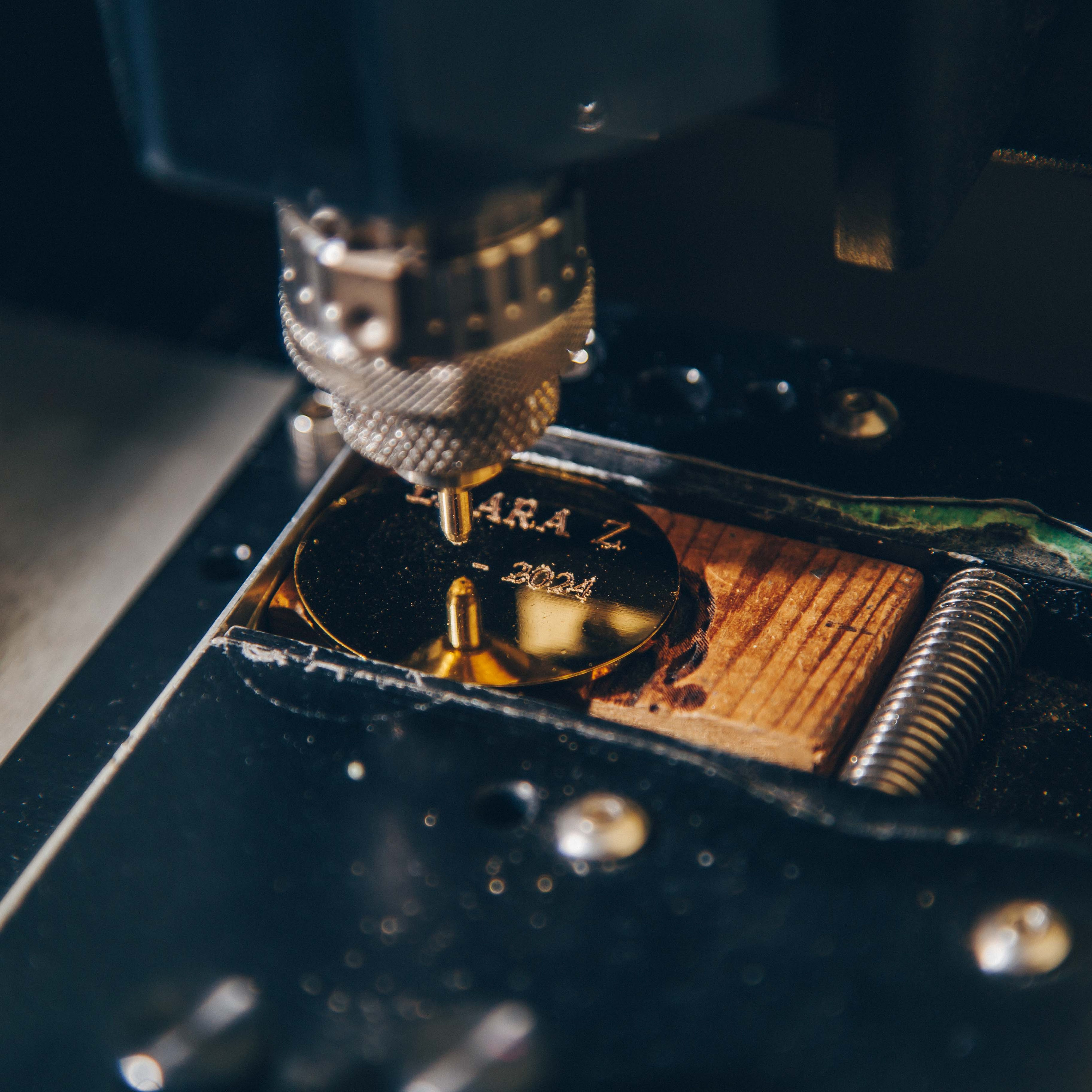 A close-up view of a metal engraving process. A precision engraving machine is actively etching text onto a shiny, circular metal plate. The text 'ARA Z' and '2024' is visible on the surface of the plate. The machine's engraving tool is positioned directly above the plate, with its metallic, textured surface in focus.