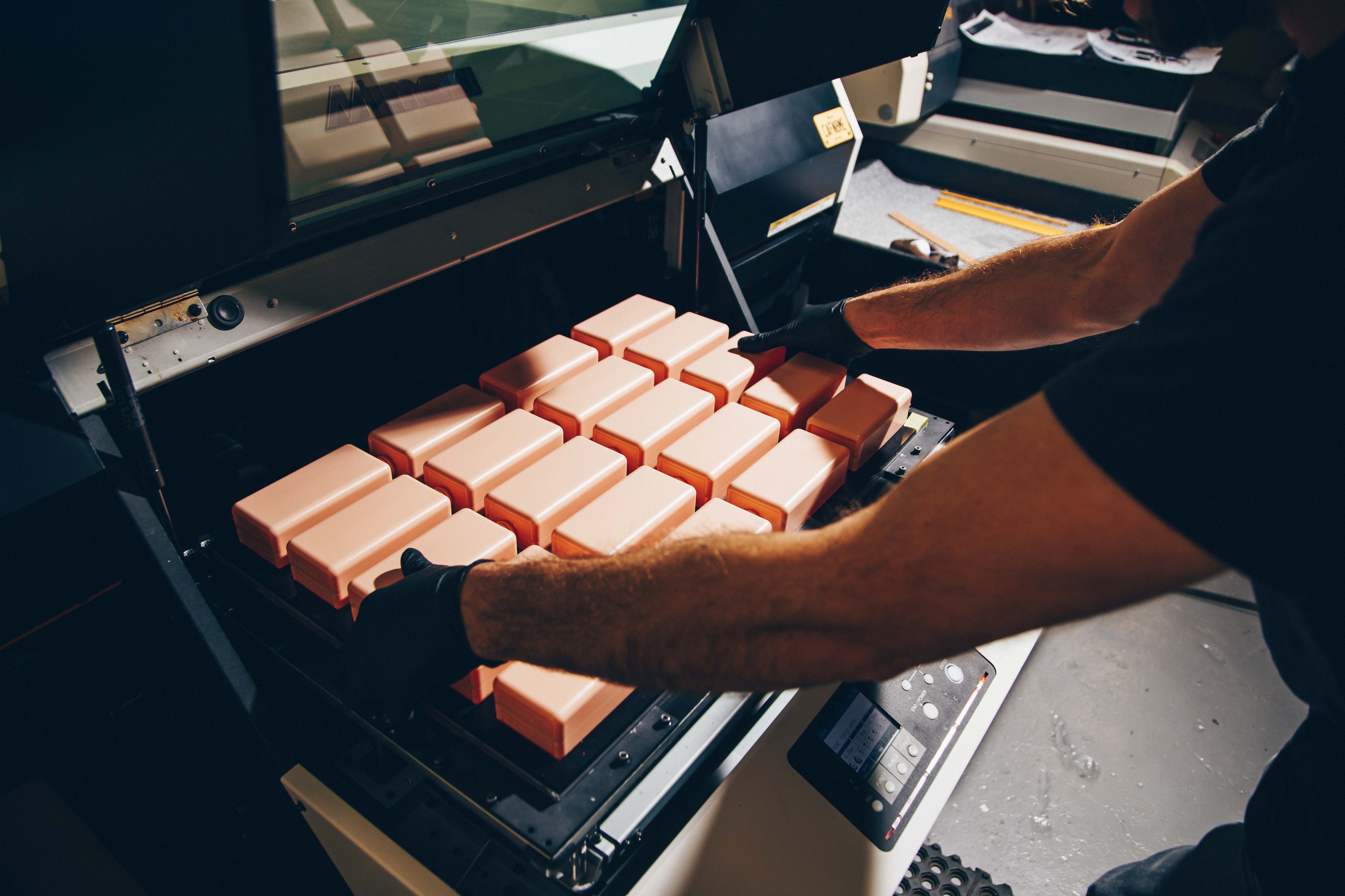A person wearing black gloves, working with a machine that appears to be a 3D printer or a similar industrial device. The person is handling a tray filled with multiple rectangular blocks, which are a light pink color. The machine is open, revealing its interior, and the blocks are neatly arranged in rows.