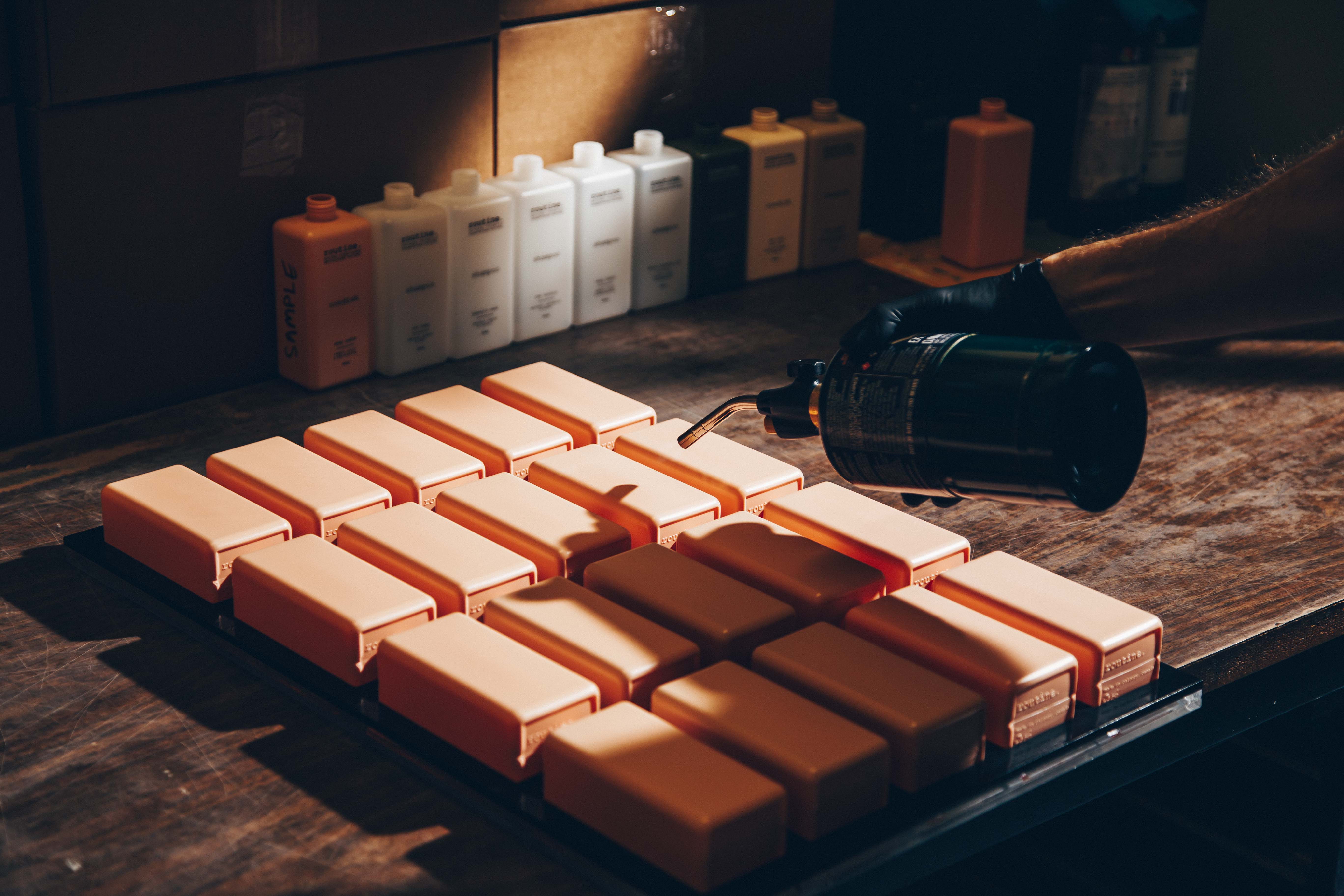 A workspace with a focus on a tray of rectangular, peach-colored shampoo bottles arranged neatly in rows. A hand wearing a black glove is holding a blowtorch, applying heat to one of the bottles. The background features a row of bottles, some labeled, lined up against a wall of cardboard boxes.