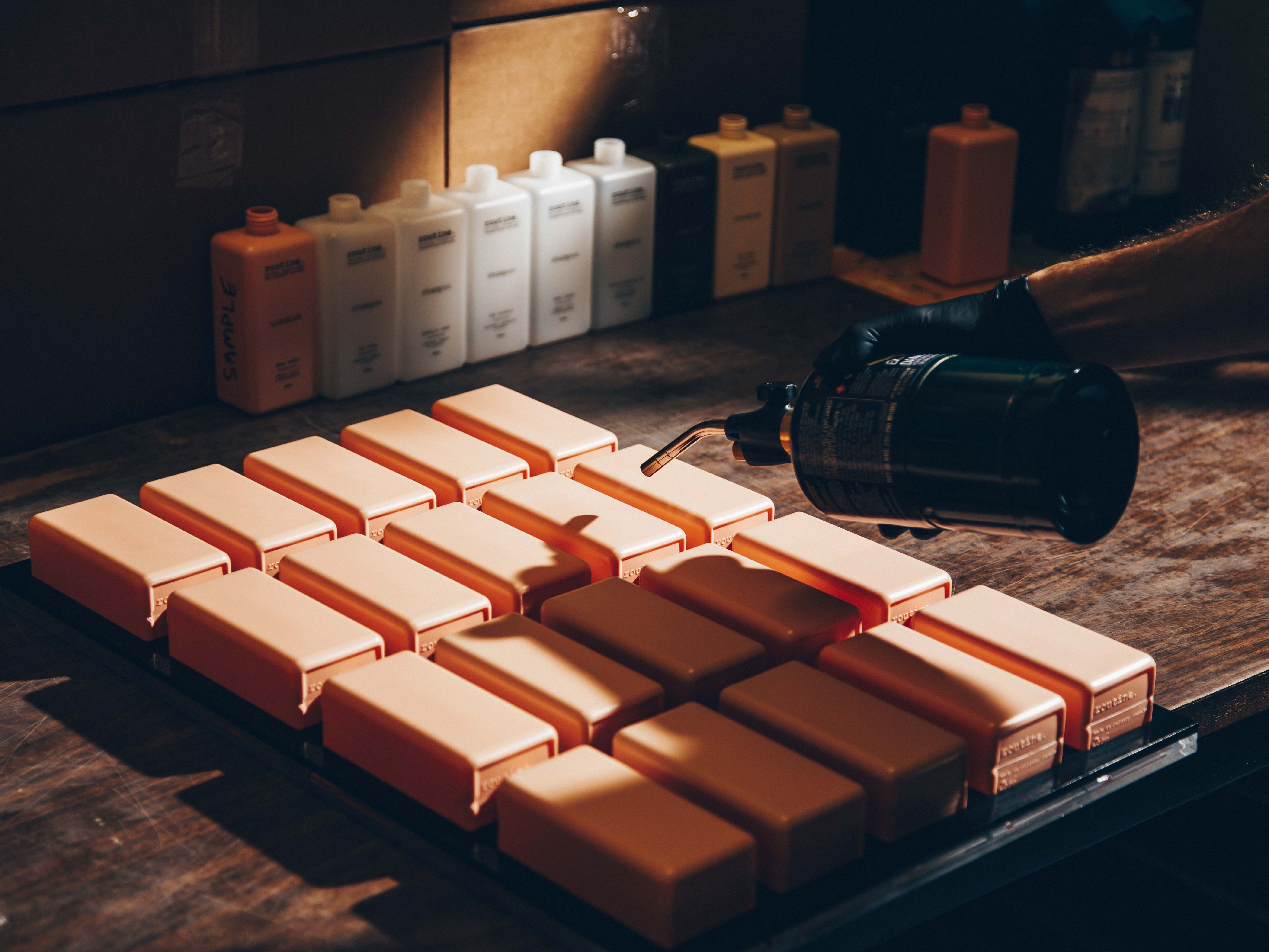 A workspace with a focus on a tray of rectangular, peach-colored shampoo bottles arranged neatly in rows. A hand wearing a black glove is holding a blowtorch, applying heat to one of the bottles. The background features a row of bottles, some labeled, lined up against a wall of cardboard boxes.