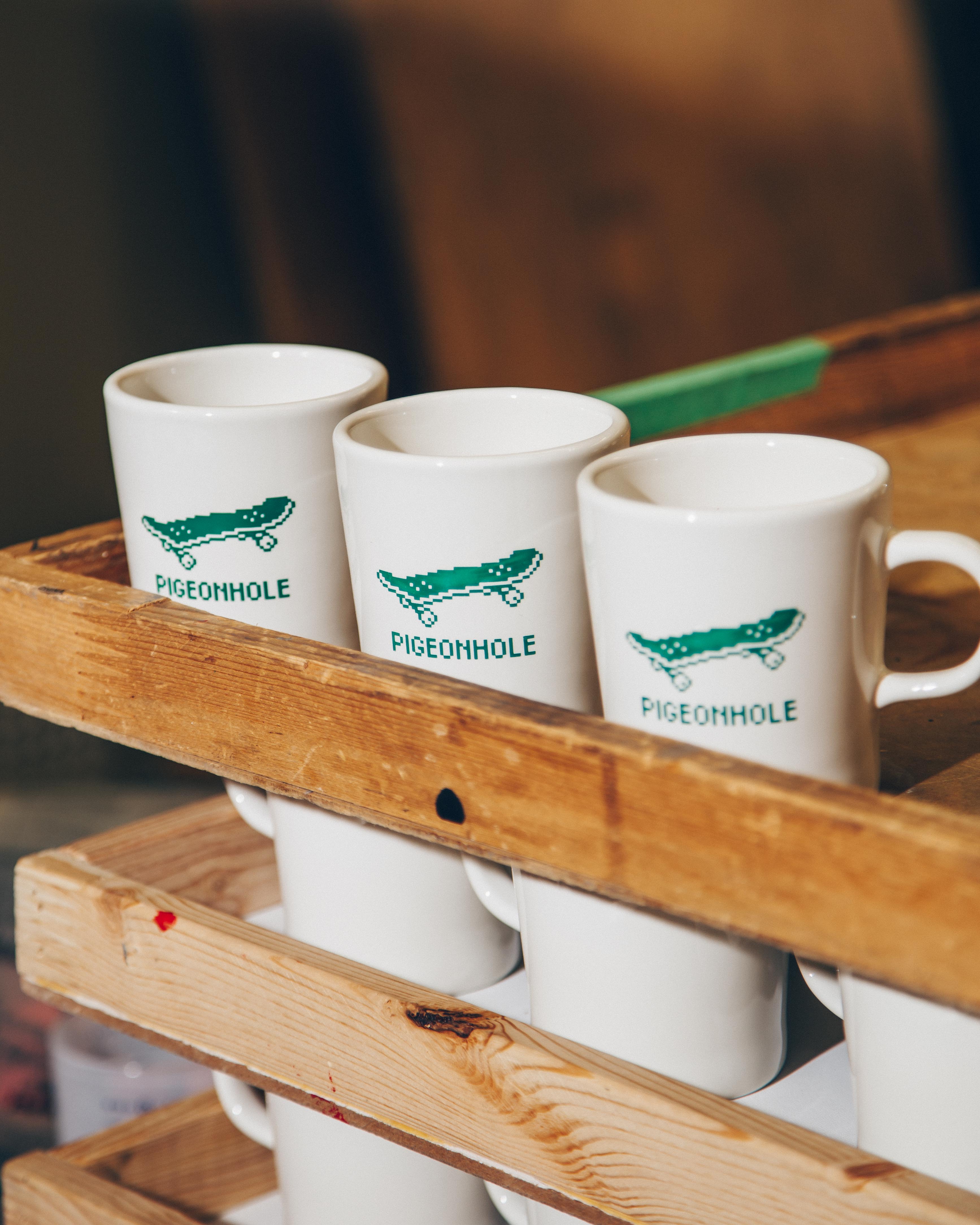 Three white mugs with green skateboard design and 'Pigeonhole' text in a wooden crate.