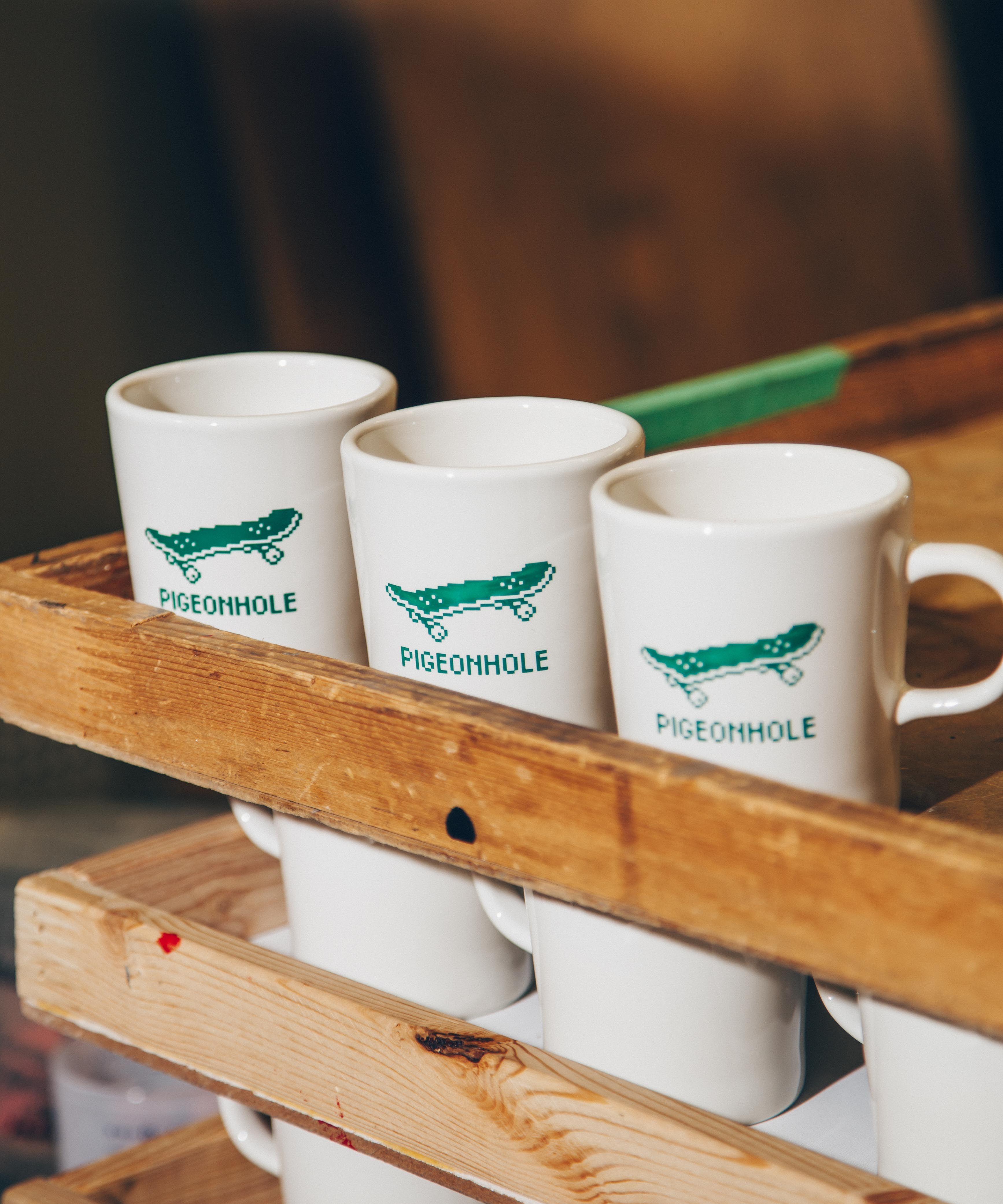 Three white mugs with green skateboard design and 'Pigeonhole' text in a wooden crate.