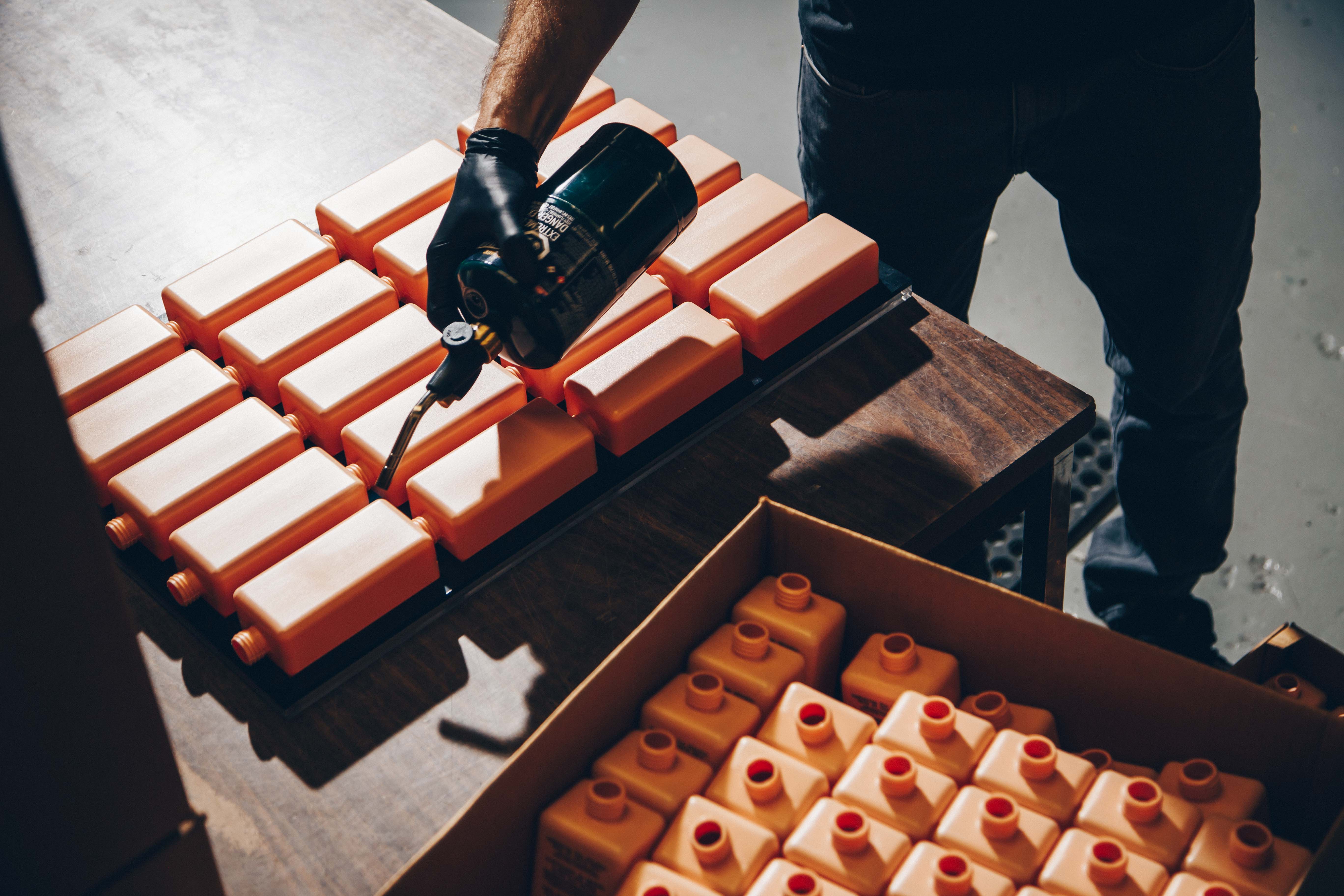 A person working with a collection of orange plastic containers. The individual is wearing a black glove and holding a propane torch, applying heat to the containers, which are neatly arranged on a wooden table.