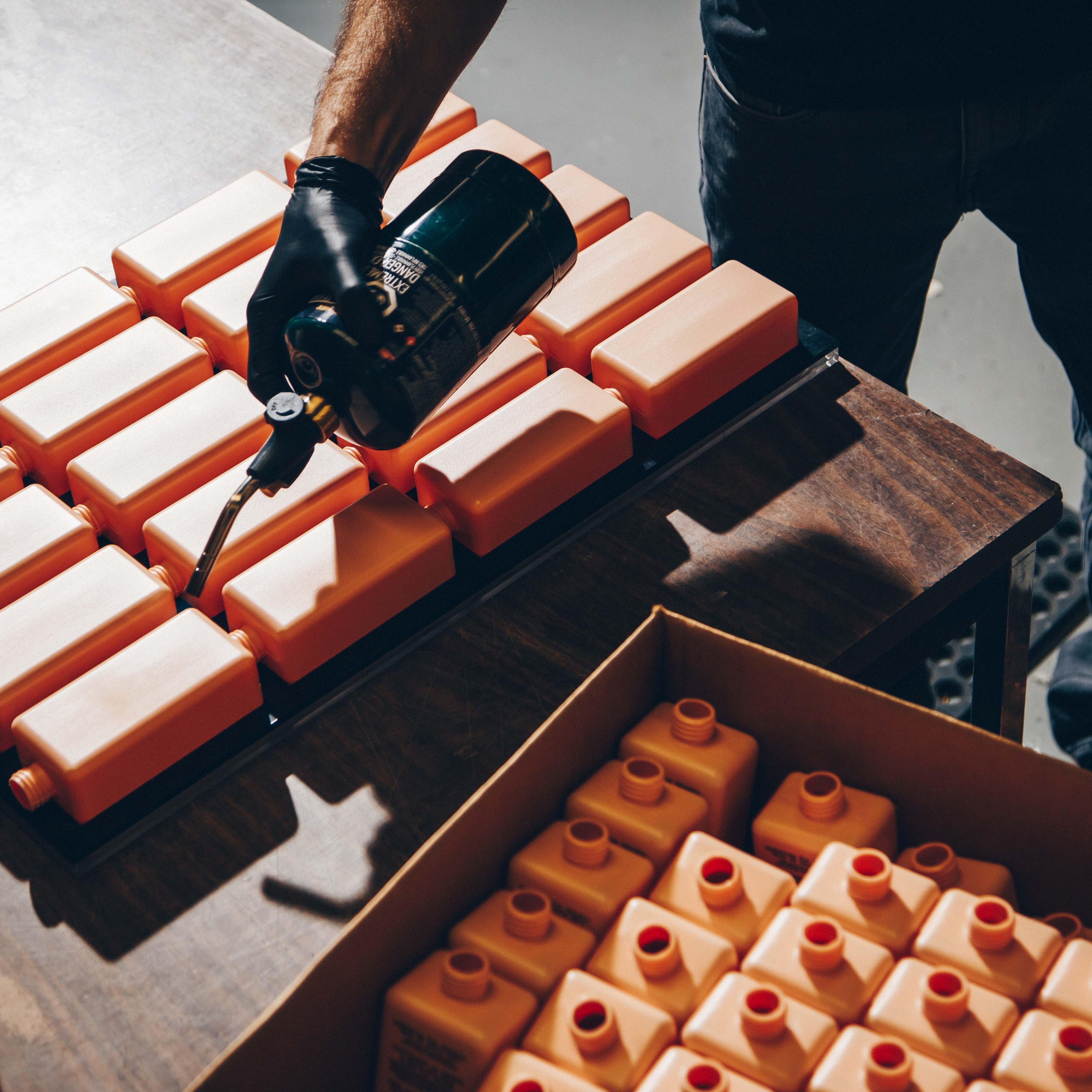 A person working with a collection of orange plastic containers. The individual is wearing a black glove and holding a propane torch, applying heat to the containers, which are neatly arranged on a wooden table.