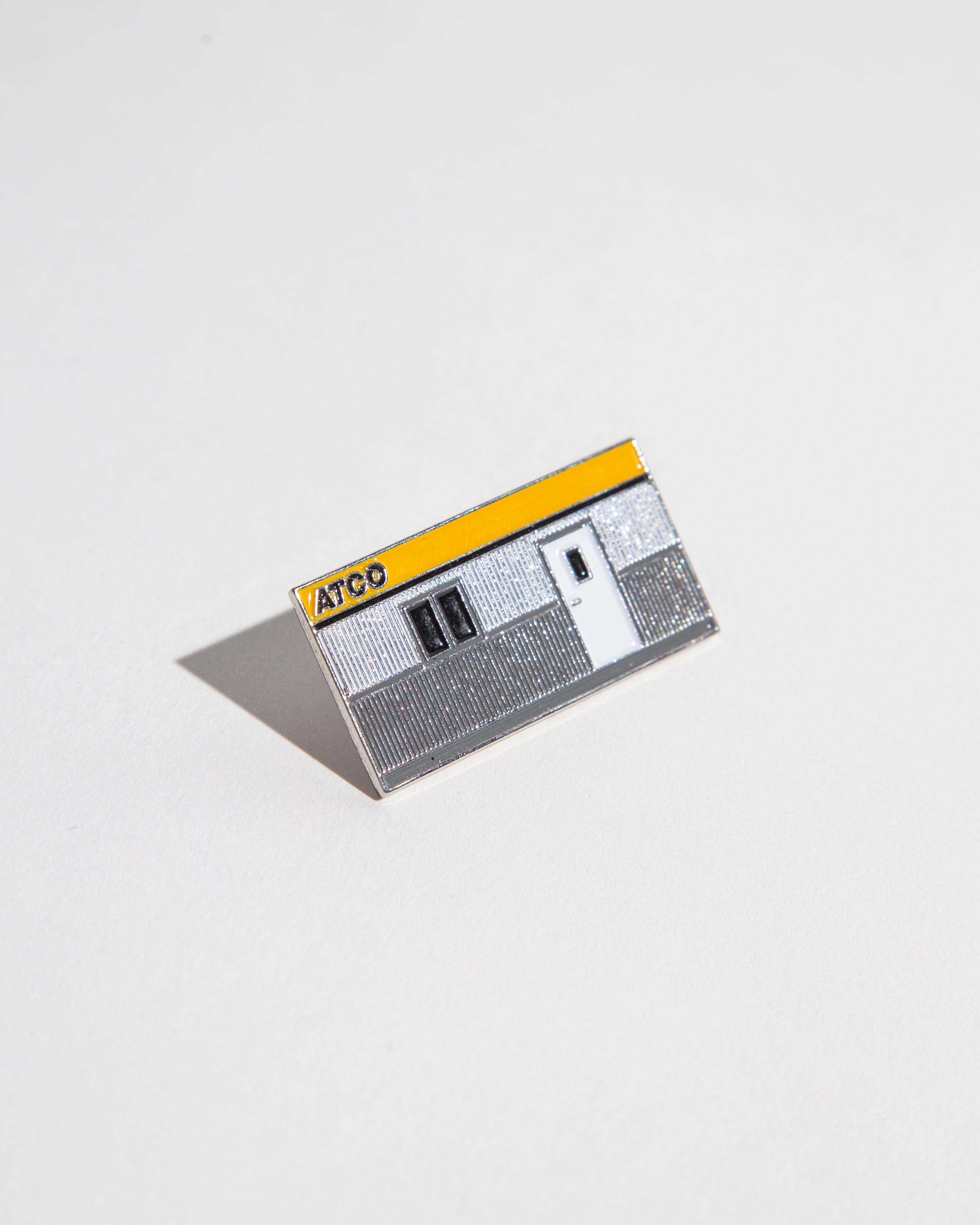 Enamel pin shaped like an ATCO building with a yellow and gray design.