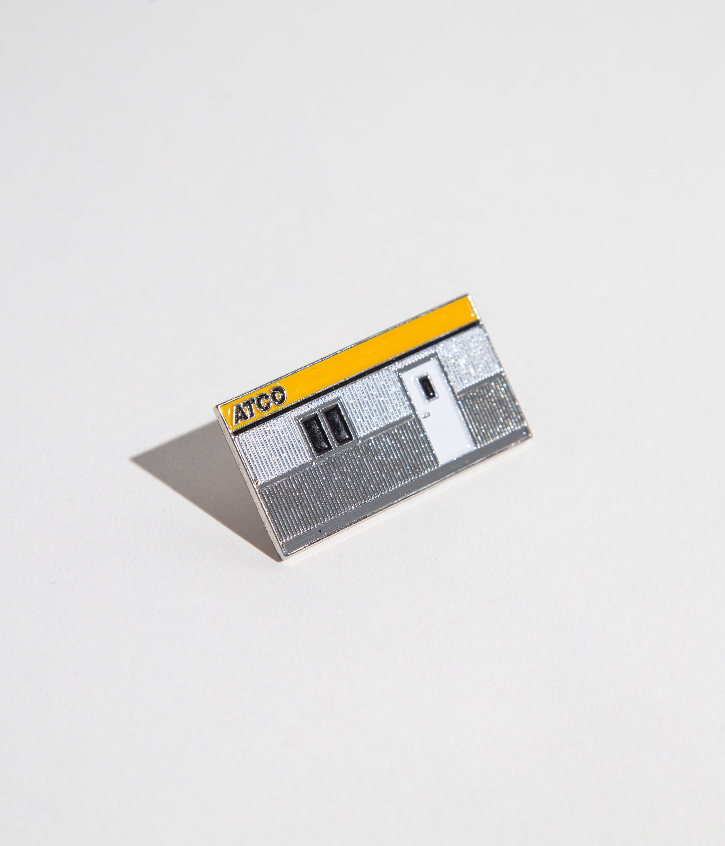 Enamel pin shaped like an ATCO building with a yellow and gray design.