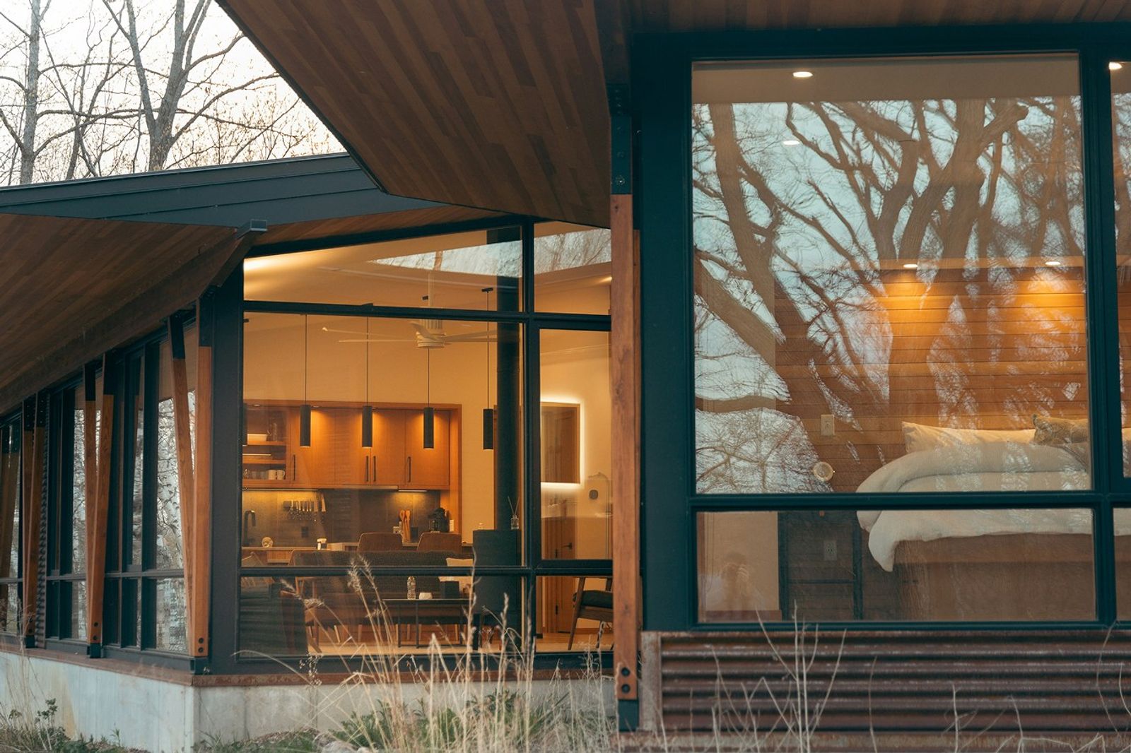 Een houten huis met grote ramen, warm licht binnen en kale bomen die weerspiegelen in het glas.
