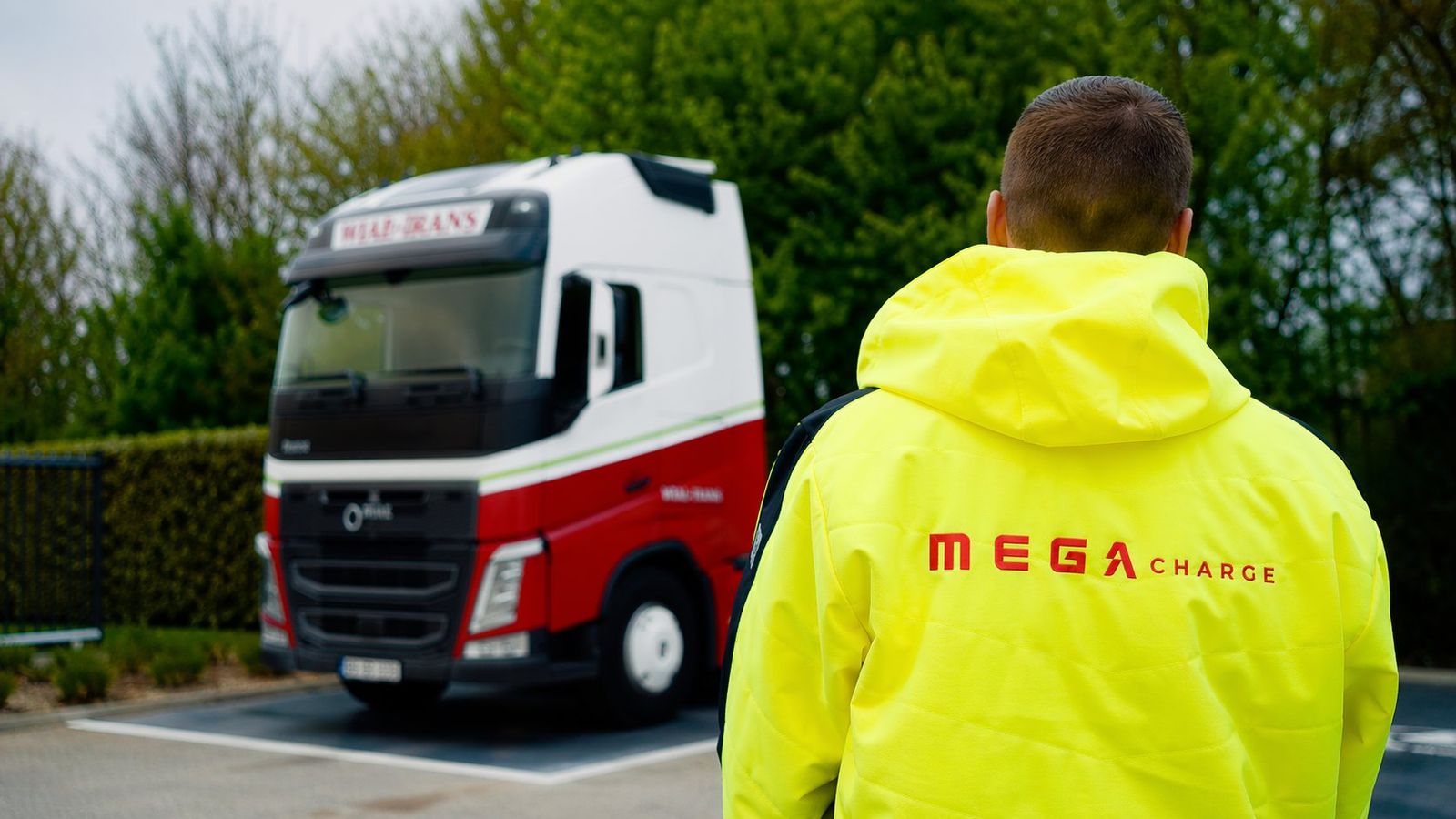 Een medewerker van MEGA Charge in een fluogele jas met het logo op de rug kijkt naar een geparkeerde vrachtwagen.