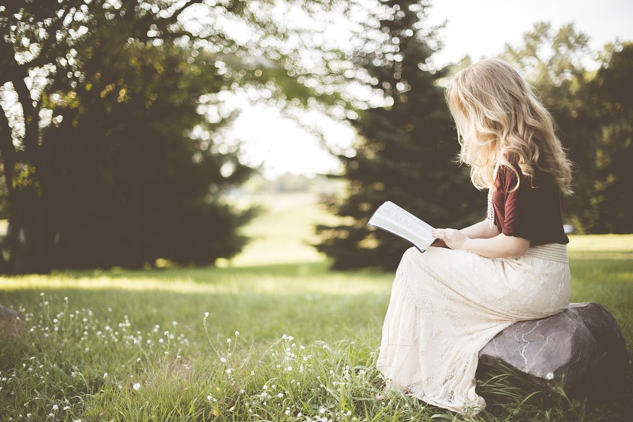 Blonde Frau liest in einem Buch, sitzt auf einer Wiese auf einem Baumstamm