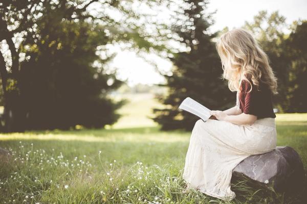 Blonde Frau liest in einem Buch, sitzt auf einer Wiese auf einem Baumstamm