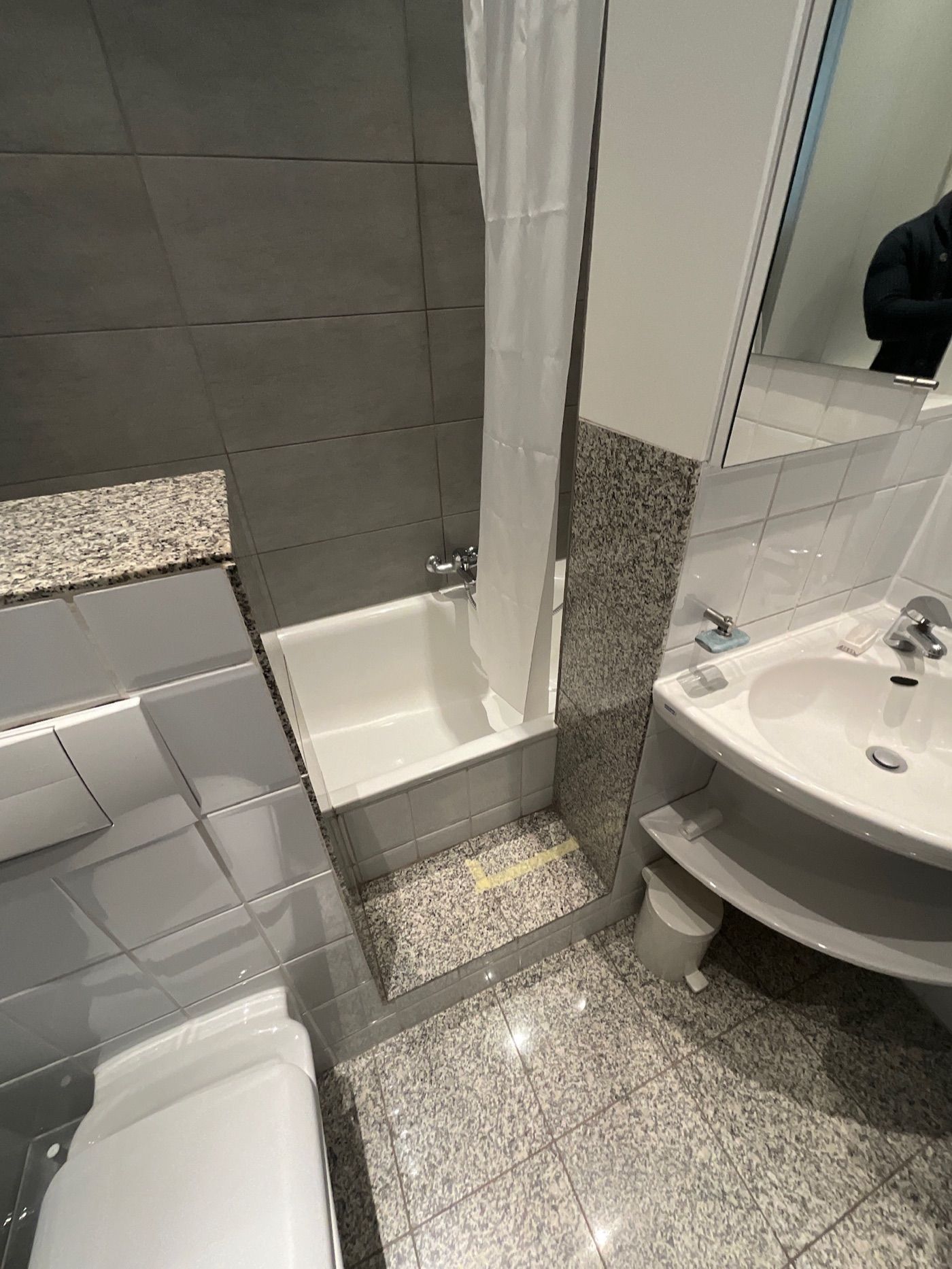 Das Badezimmer vor der Sanierung: Badewanne mit Duschvorhang, Eckwaschbecken und Bodenfliesen in Granitoptik.