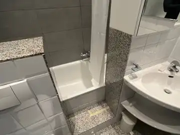 Das Badezimmer vor der Sanierung: Badewanne mit Duschvorhang, Eckwaschbecken und Bodenfliesen in Granitoptik.