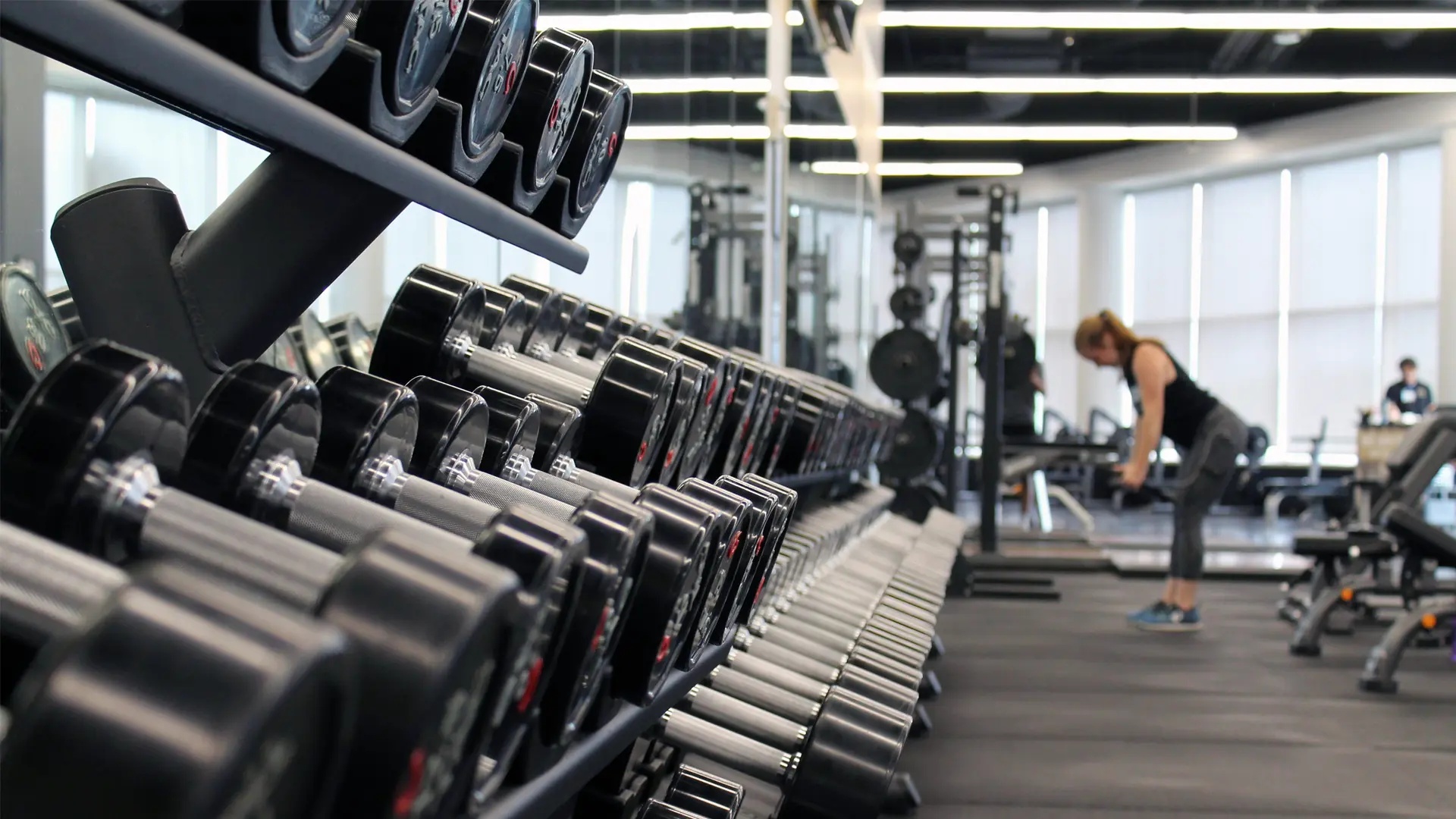 Bild på vikter på ett gym. En person som tränar syns i bakgrunden.