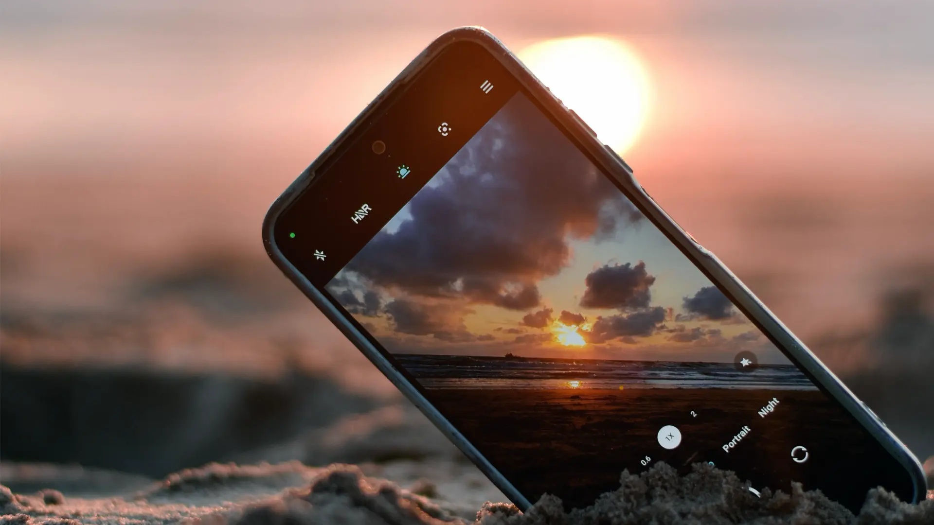 Mobiltelefon på sandstrand fotograferar solnedgång.