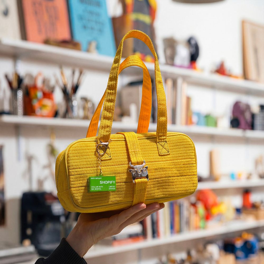 Nik's hand holds the yellow shipping strap bag in the air. the Studio's wall of past and present products is blurry in the background.