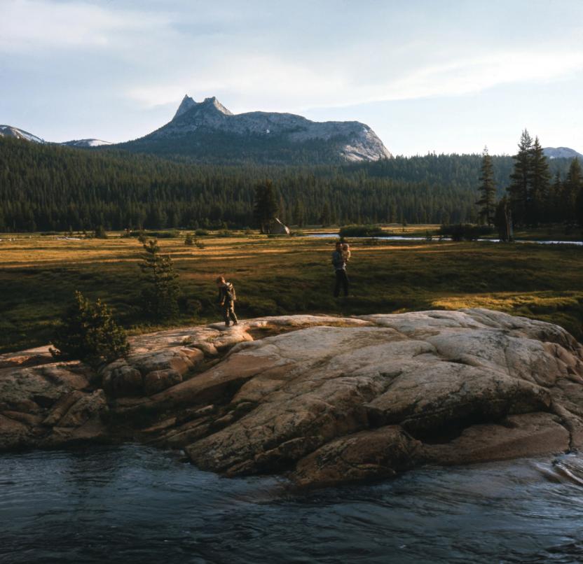 two people are standing on a rock near a river with mountains in the background .