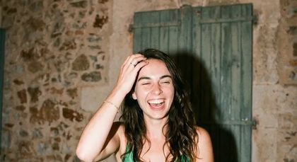 Une invitée en robe verte éclate de rire, la main dans les cheveux, devant un mur de pierre.