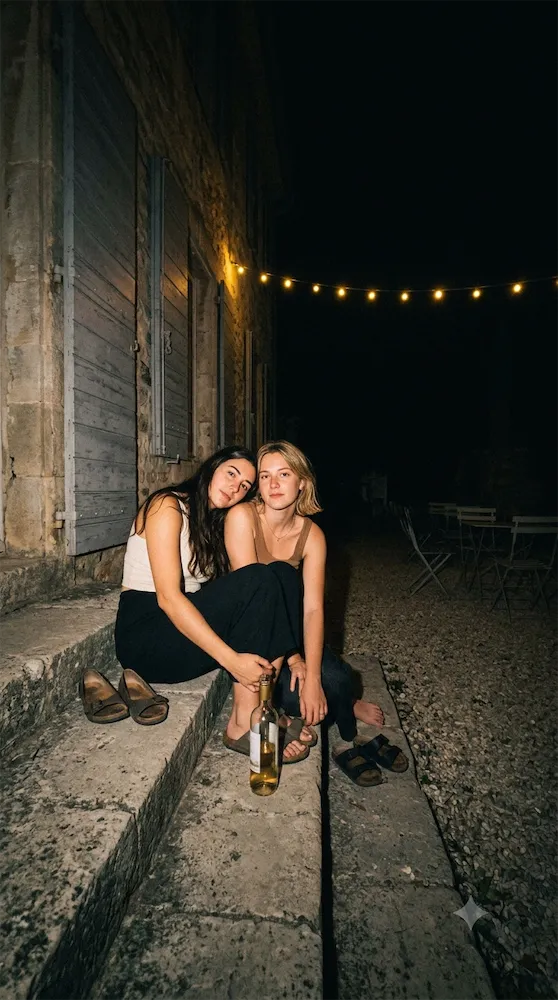 Deux amies assises sur les marches dans la nuit, chaussures enlevées, une bouteille entre elles.