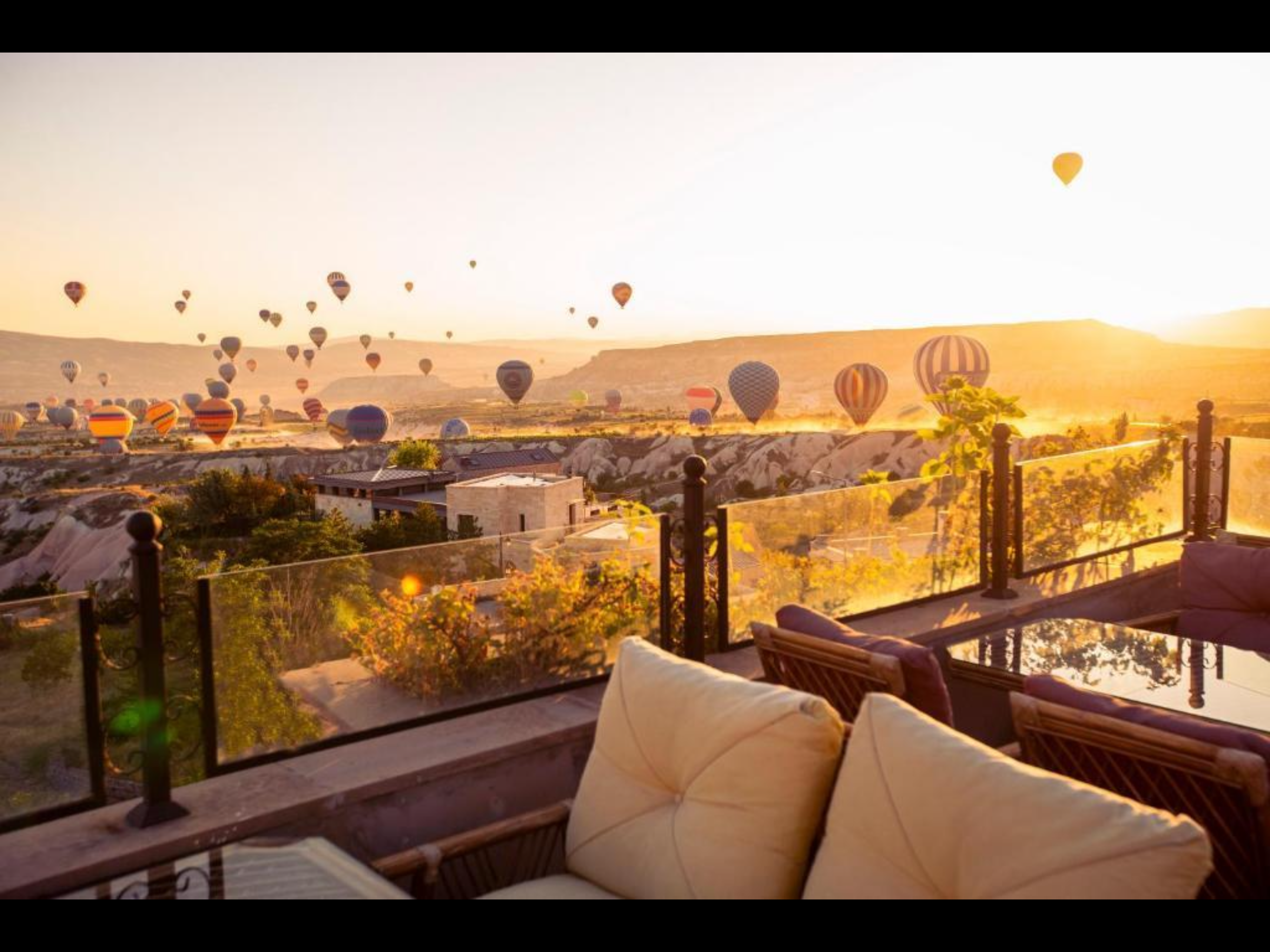 Hot air balloons float over a valley at sunrise, seen from a terrace.
