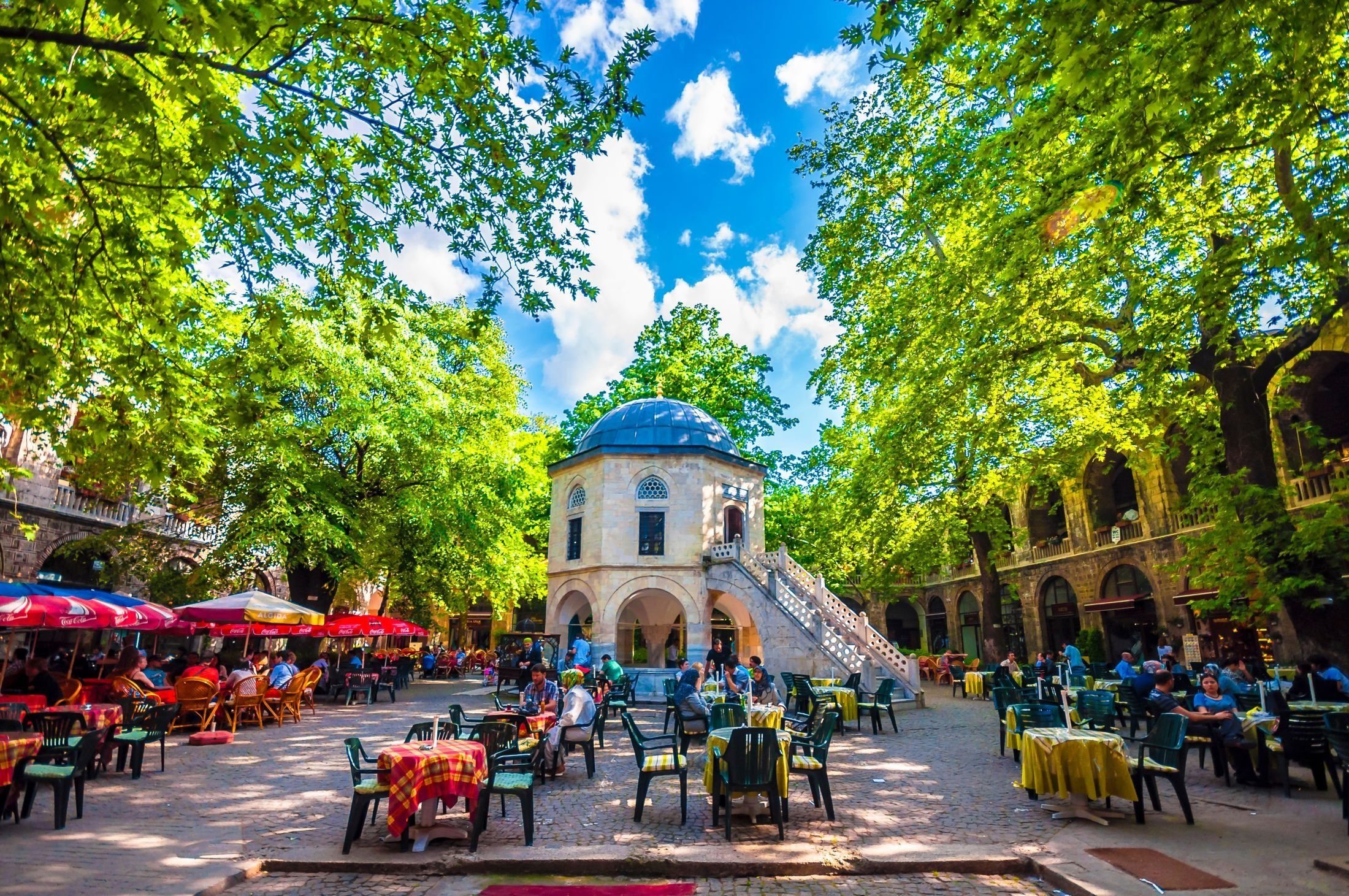 Historic courtyard with a domed pavilion, bustling outdoor cafes, and lush trees.