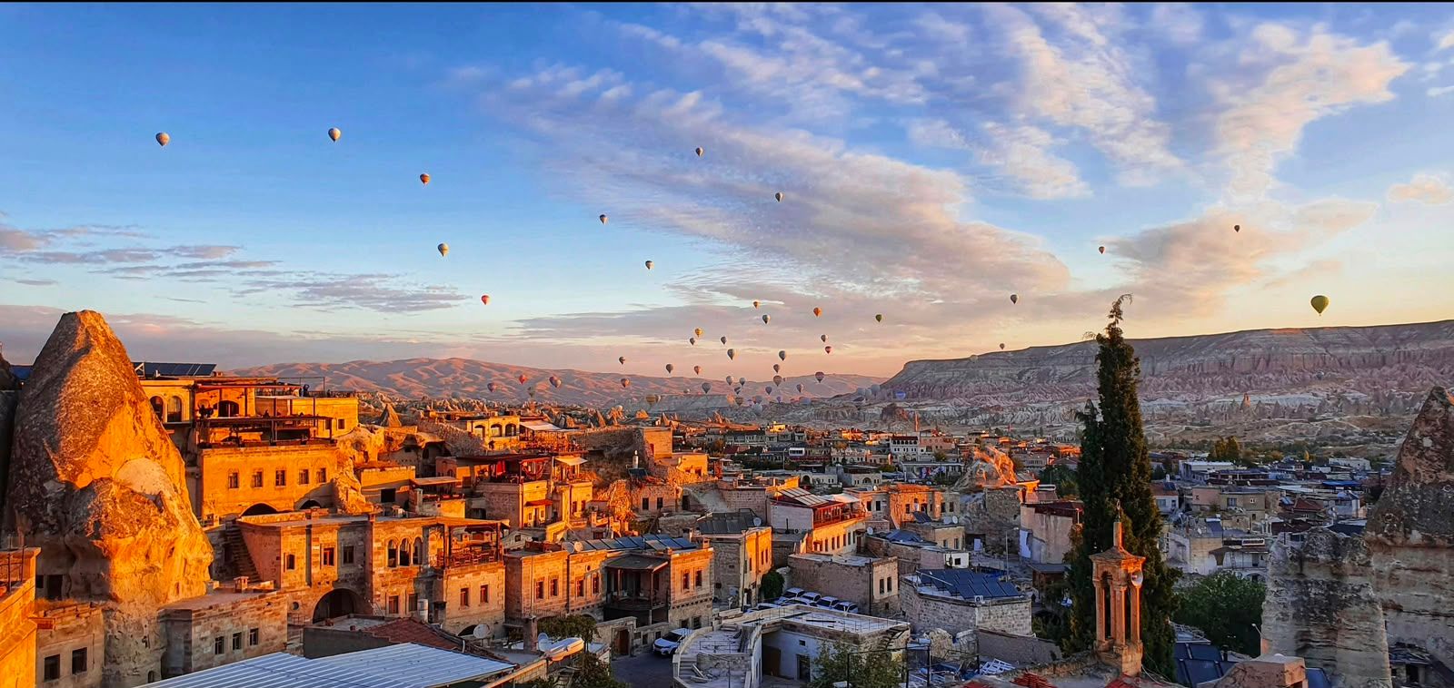 Dozens of hot air balloons float over a stone town at golden sunrise.