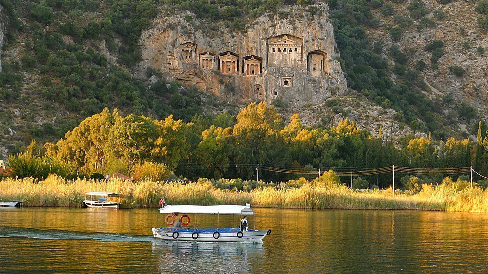 Ancient rock-cut tombs carved into a cliff face above a river with a passing boat.