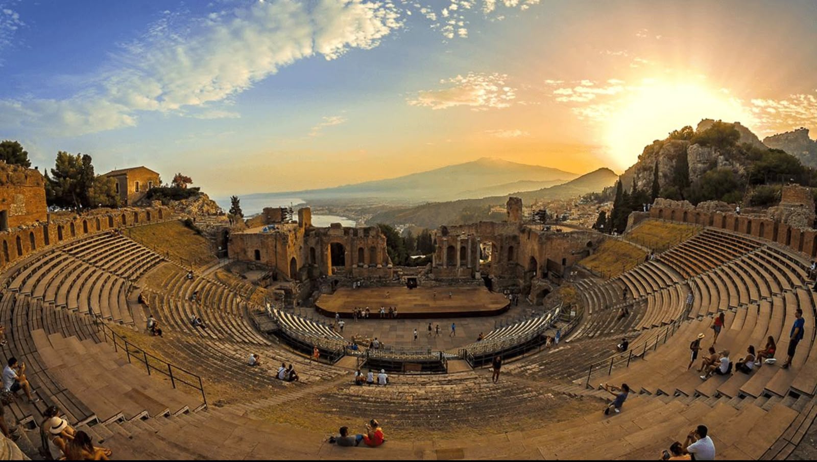 An ancient stone amphitheater under a warm orange sunset with mountains in the background.