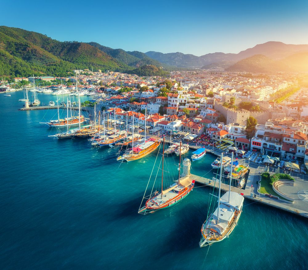 Aerial view of a bustling coastal town with a marina, historic buildings, and green mountains.