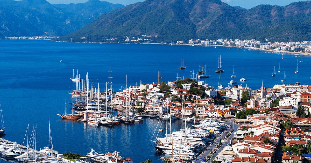 A scenic harbor town with yachts, blue sea, and green mountains.